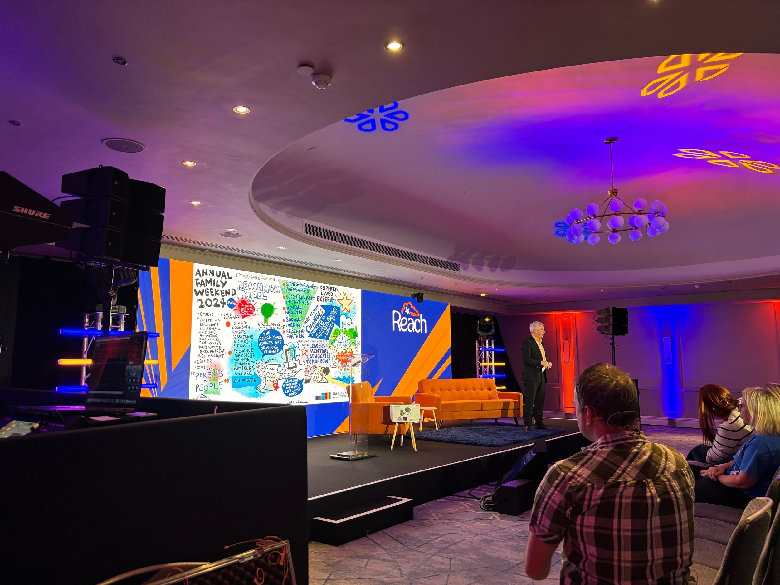 Indoor conference room with a speaker on stage, a large colorful presentation slide, and an audience. The room has blue and orange lighting accents on the ceiling and walls.
