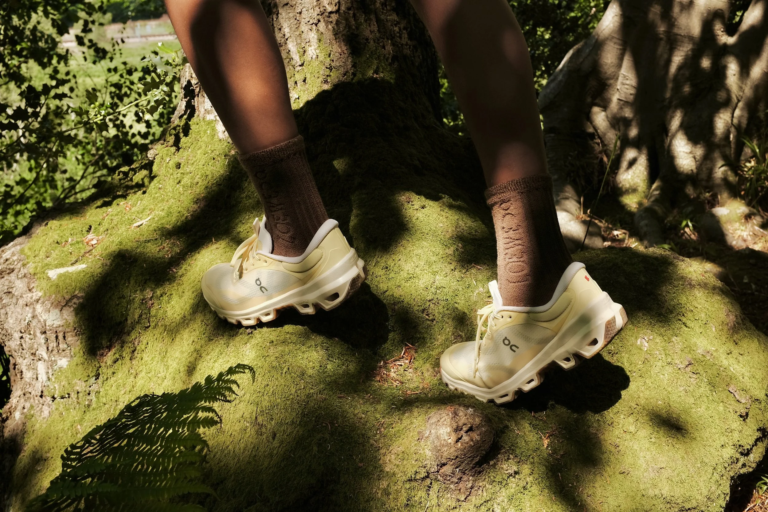 Close-up of a person's legs wearing brown socks and yellow sneakers, standing on a moss-covered tree trunk in a lush green forest.