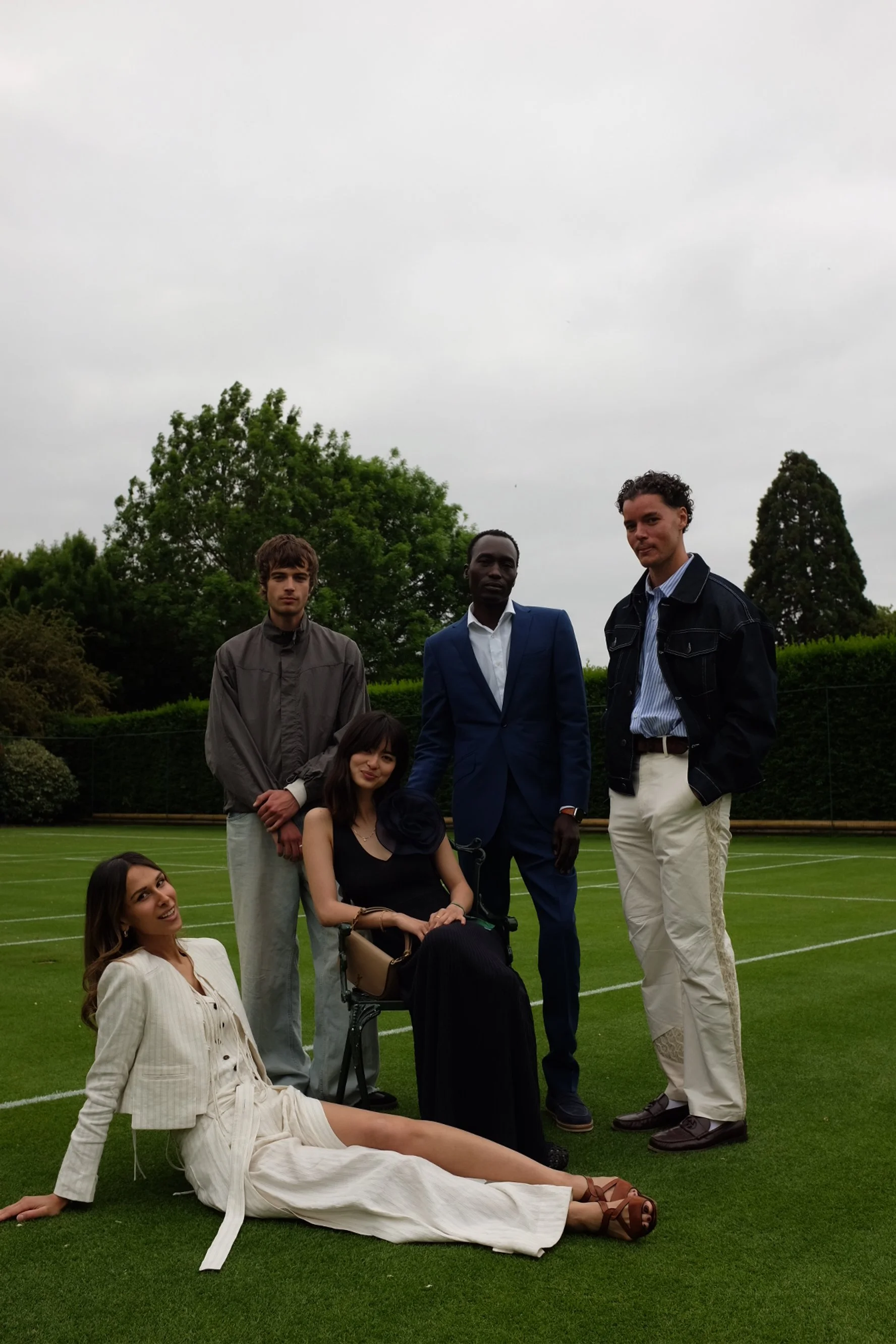 Group of five diverse young adults outdoors on a grassy field with trees in the background. Two women are seated or lying on the grass and three men are standing behind them, all dressed in stylish, semi-formal clothing.