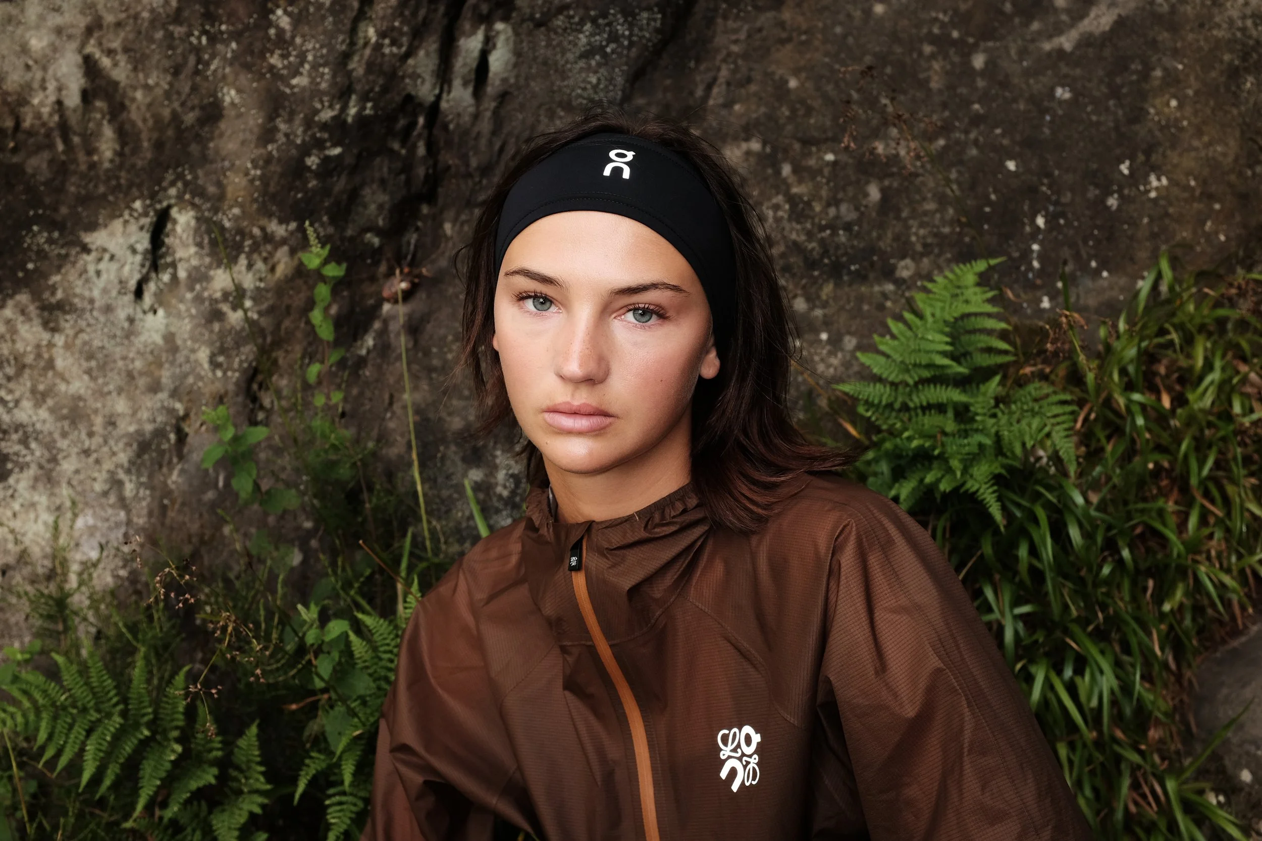 A young woman with brown hair and blue eyes wearing a black headband and brown outdoor jacket, sitting in front of a rocky background with green ferns and plants.