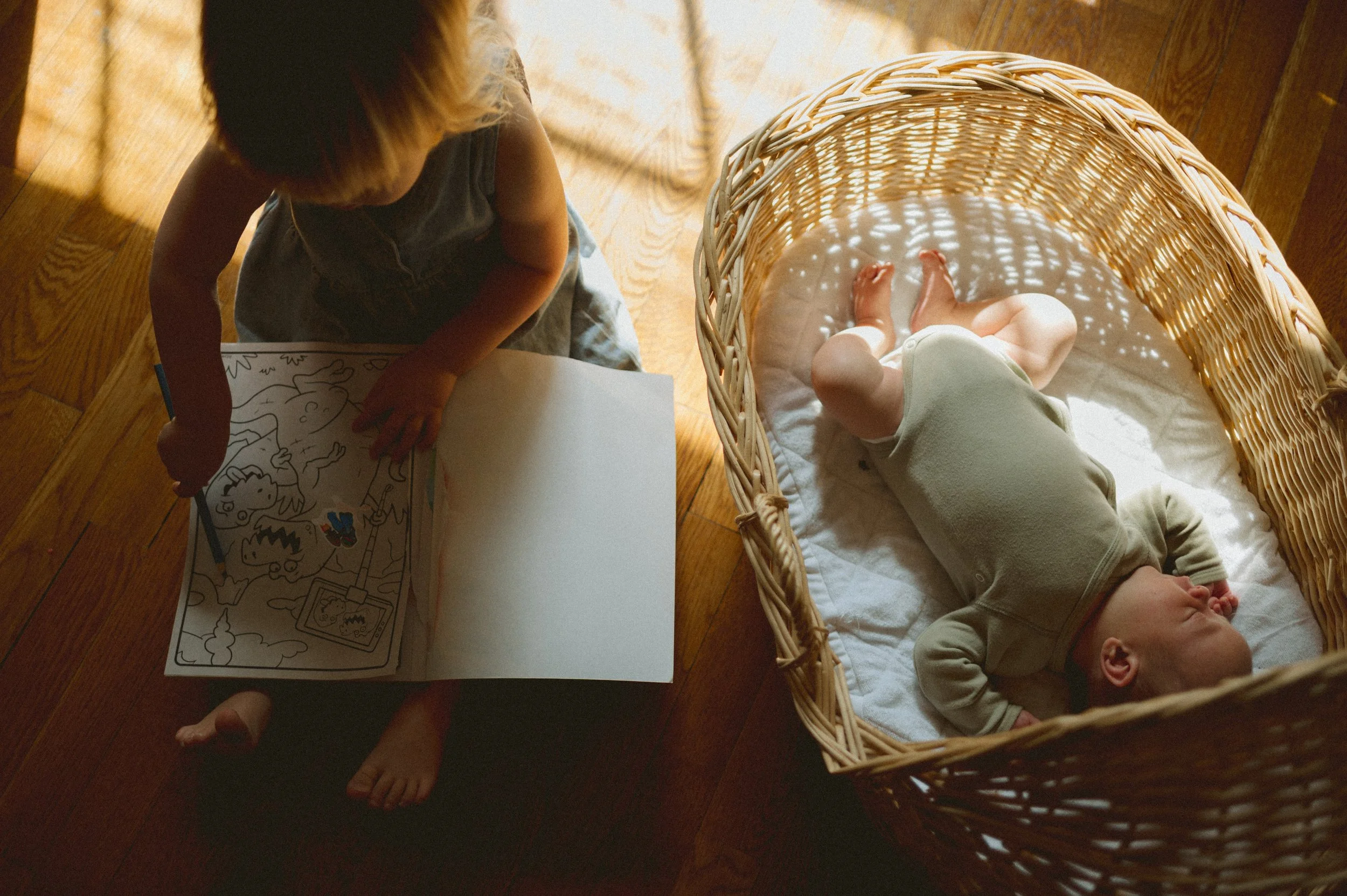 A young girl drawing with crayons at a table, beside a baby sleeping in a wicker cradle on a quilted blanket.