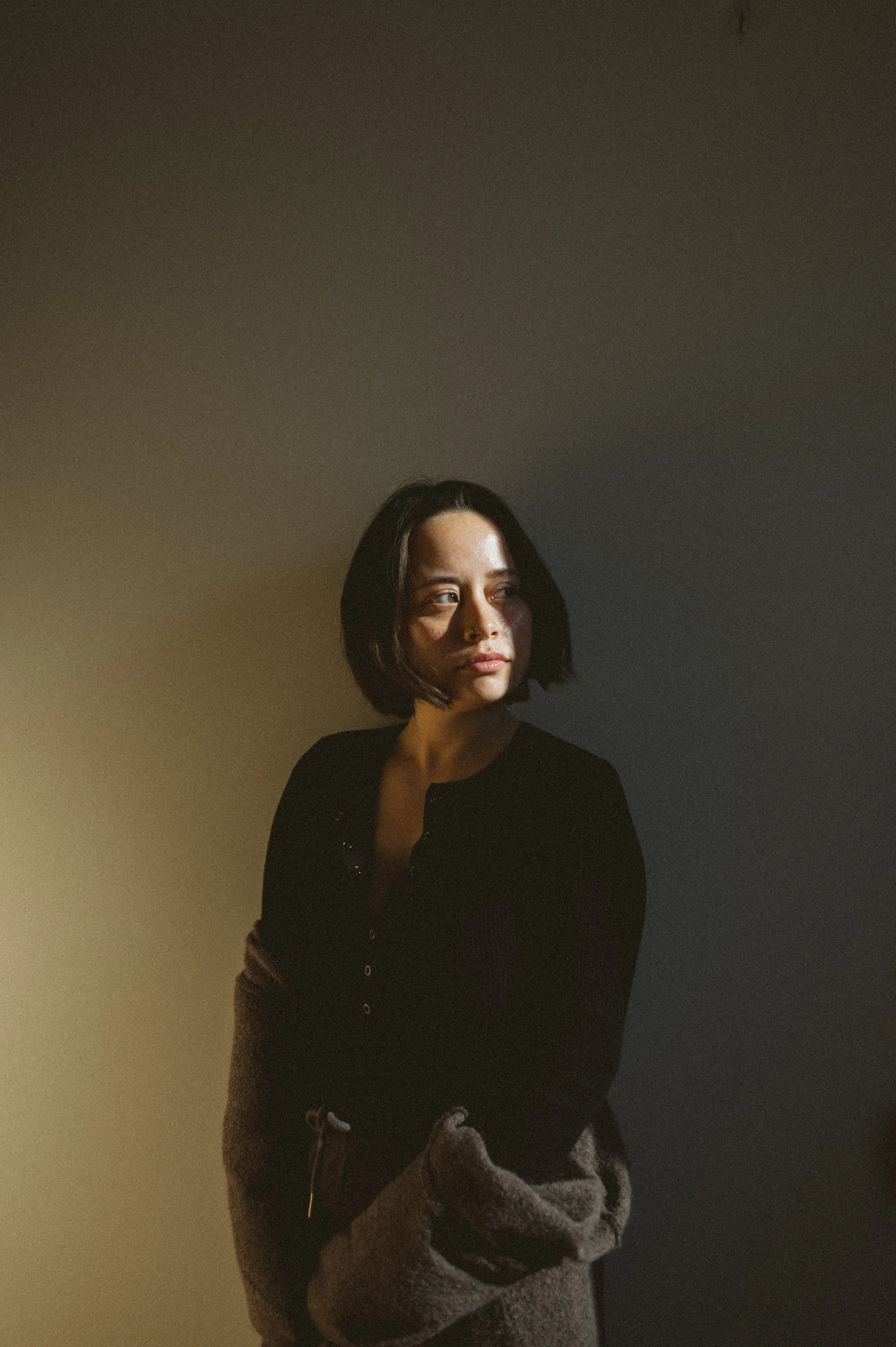 A woman with shoulder-length dark hair looking to the side, wearing a black top and gray pants, standing in a dimly lit room with shadows and subdued lighting.