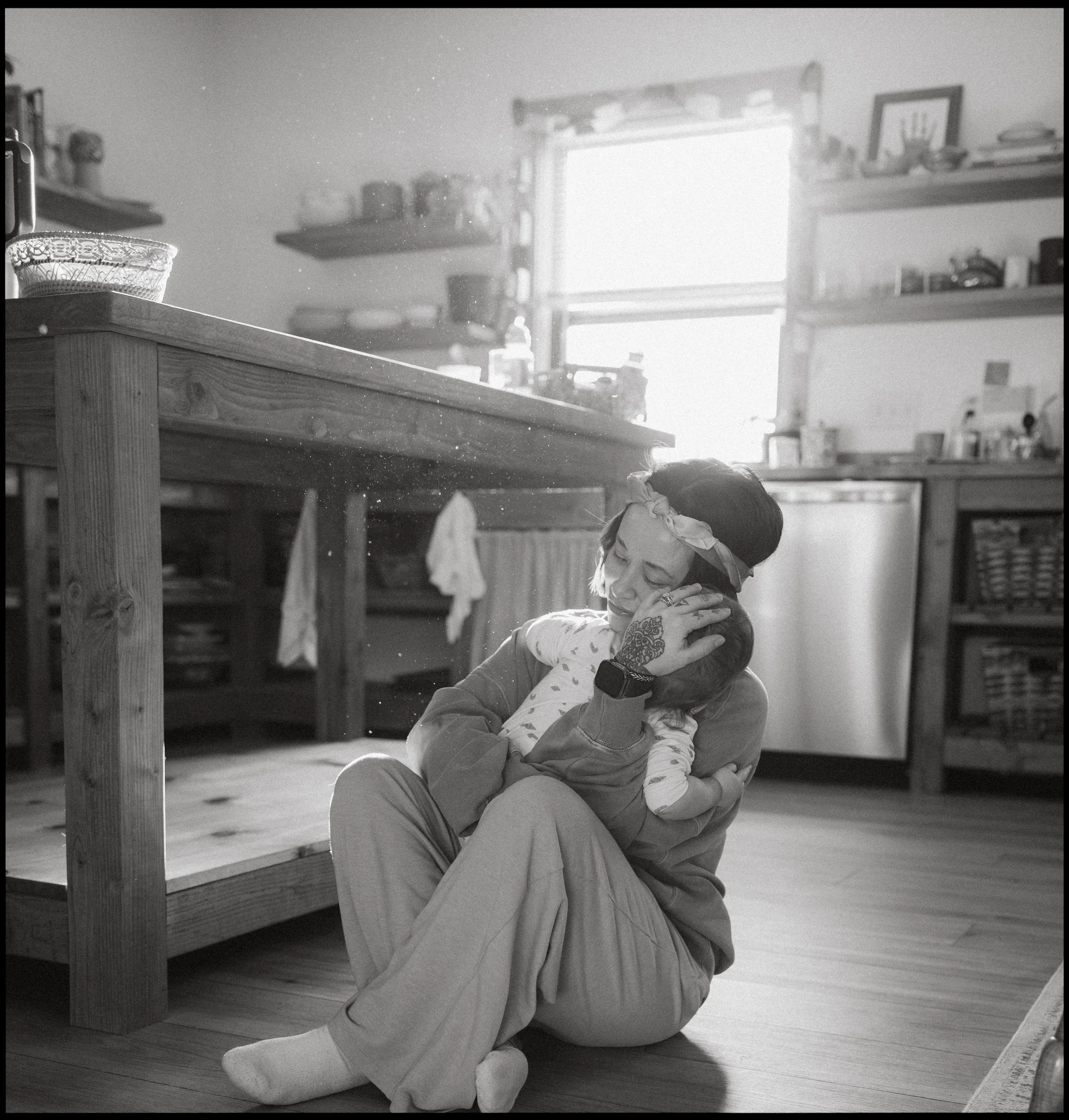 A woman with a headband holding a child in a kitchen, sitting on the floor and smiling while embracing the child.