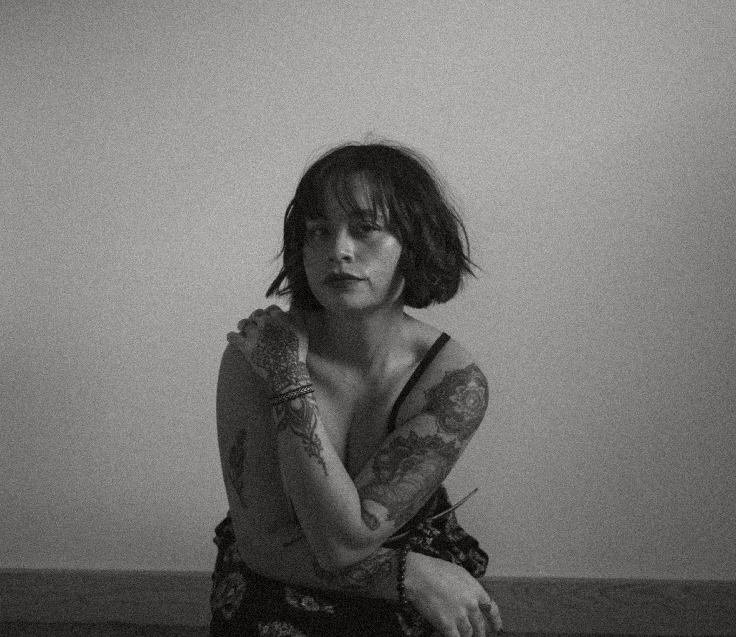 A black and white photo of a woman with short, wavy hair and tattoos on her arms and shoulder, sitting cross-legged against a plain wall.