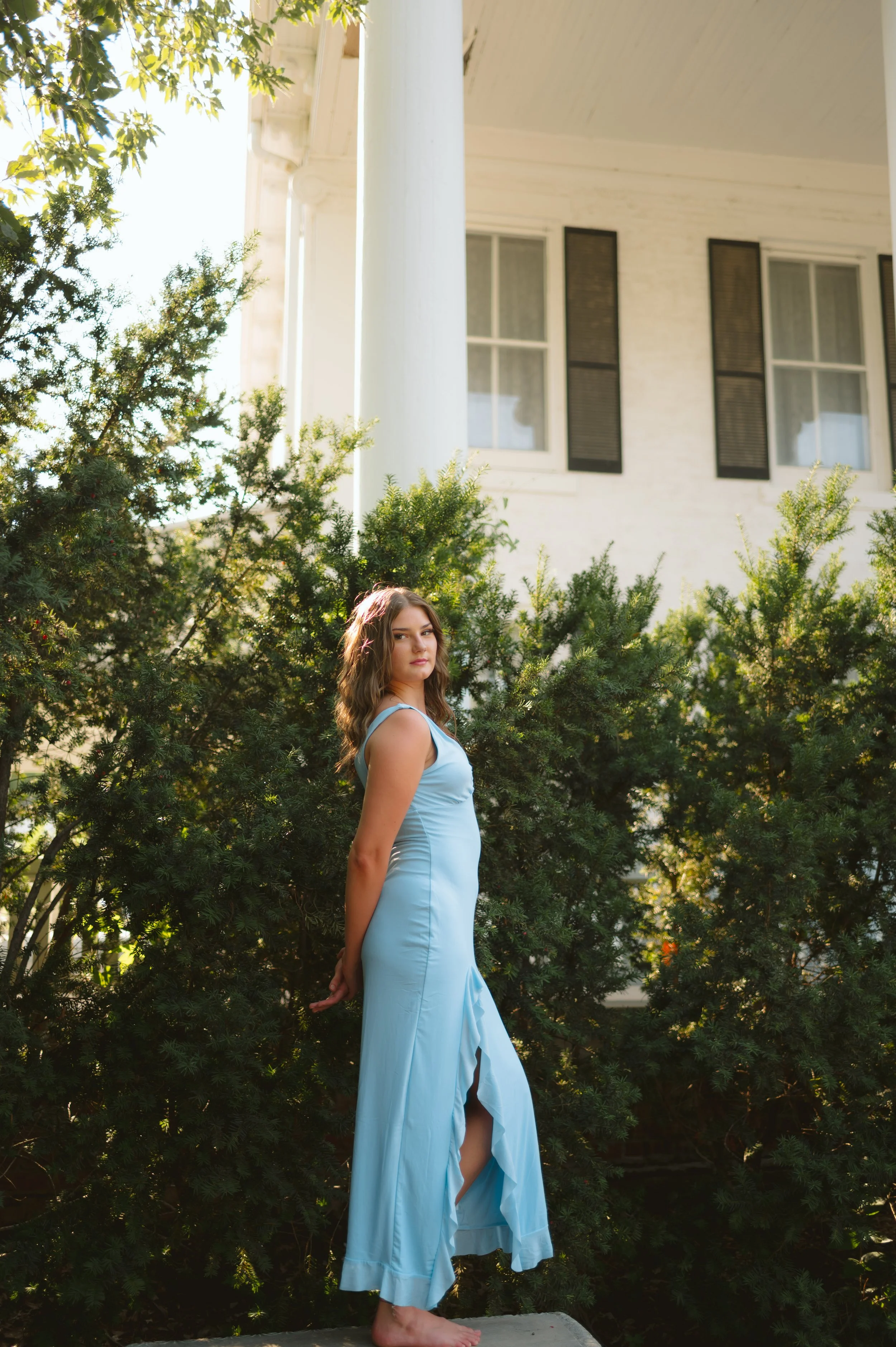A young woman with wavy brown hair wearing a light blue sleeveless dress stands barefoot on a concrete slab in front of lush green bushes, with a white house featuring black shutters in the background.