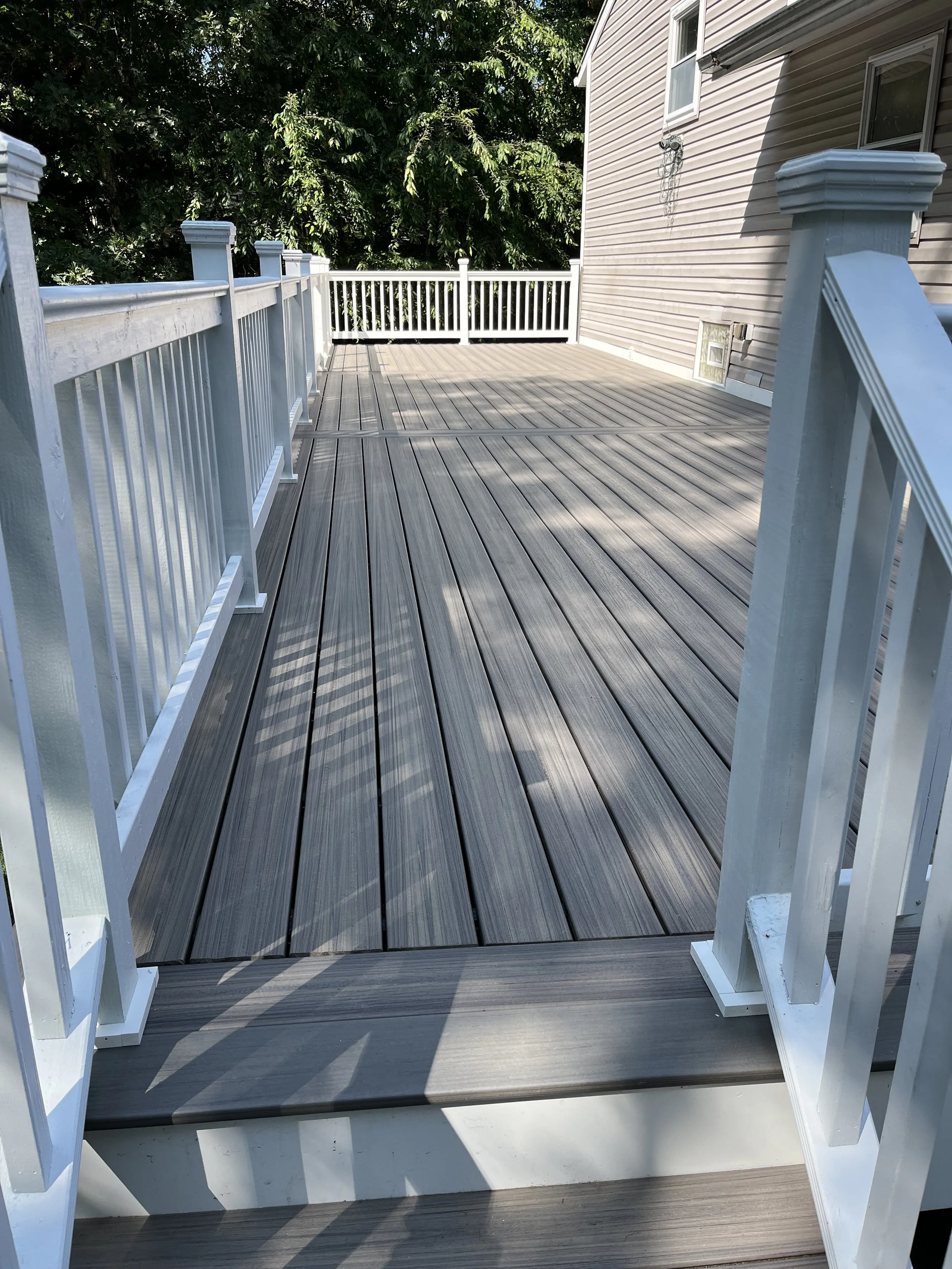 A spacious wooden deck attached to a house with siding, surrounded by a white railing, with trees in the background.