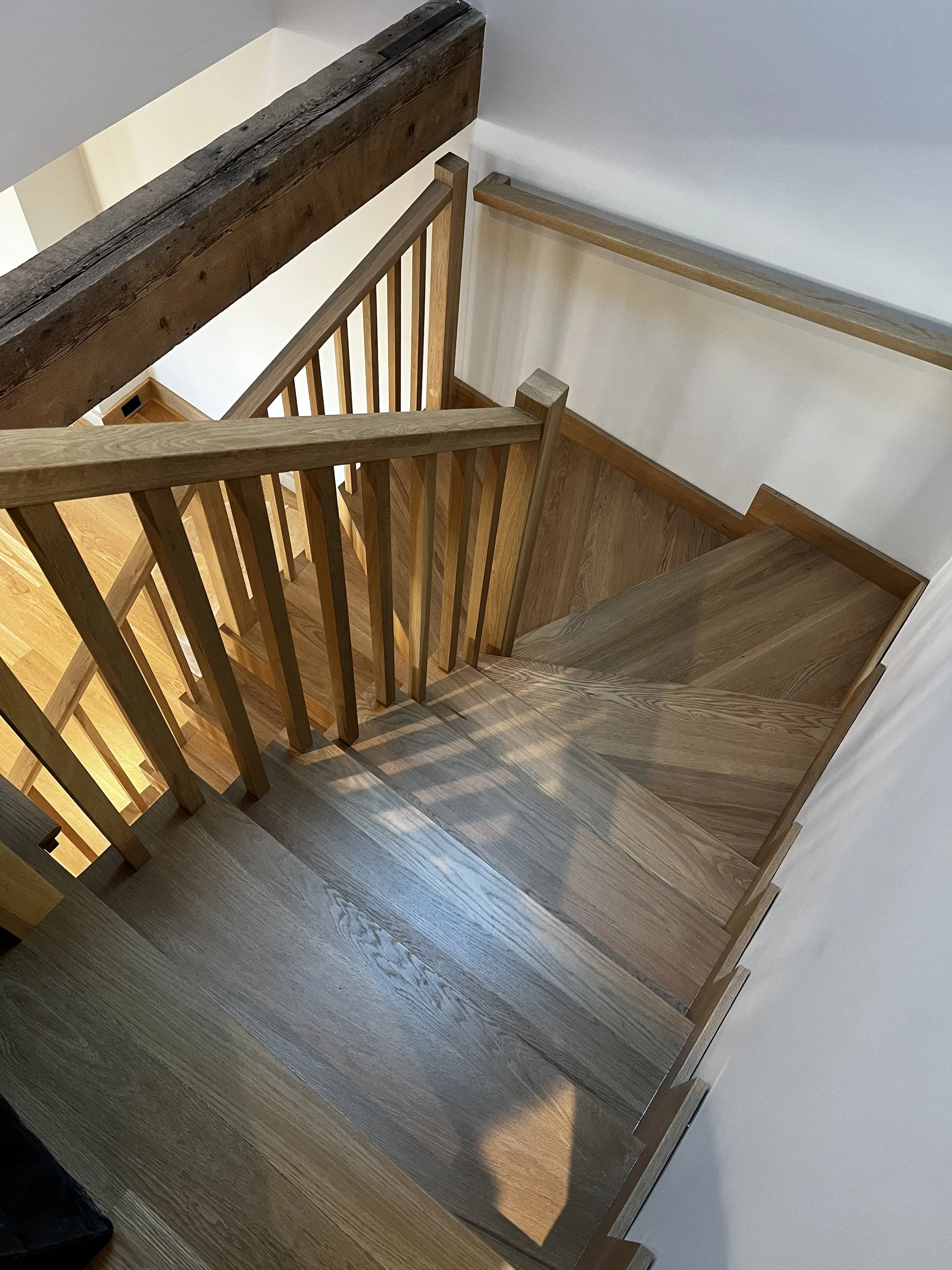 Wooden staircase with railings leading down to a lower floor, seen from the top. The stairs and railings are made of natural wood, with some darker wood accents. The view looks down from an upper level, with white walls surrounding the staircase.