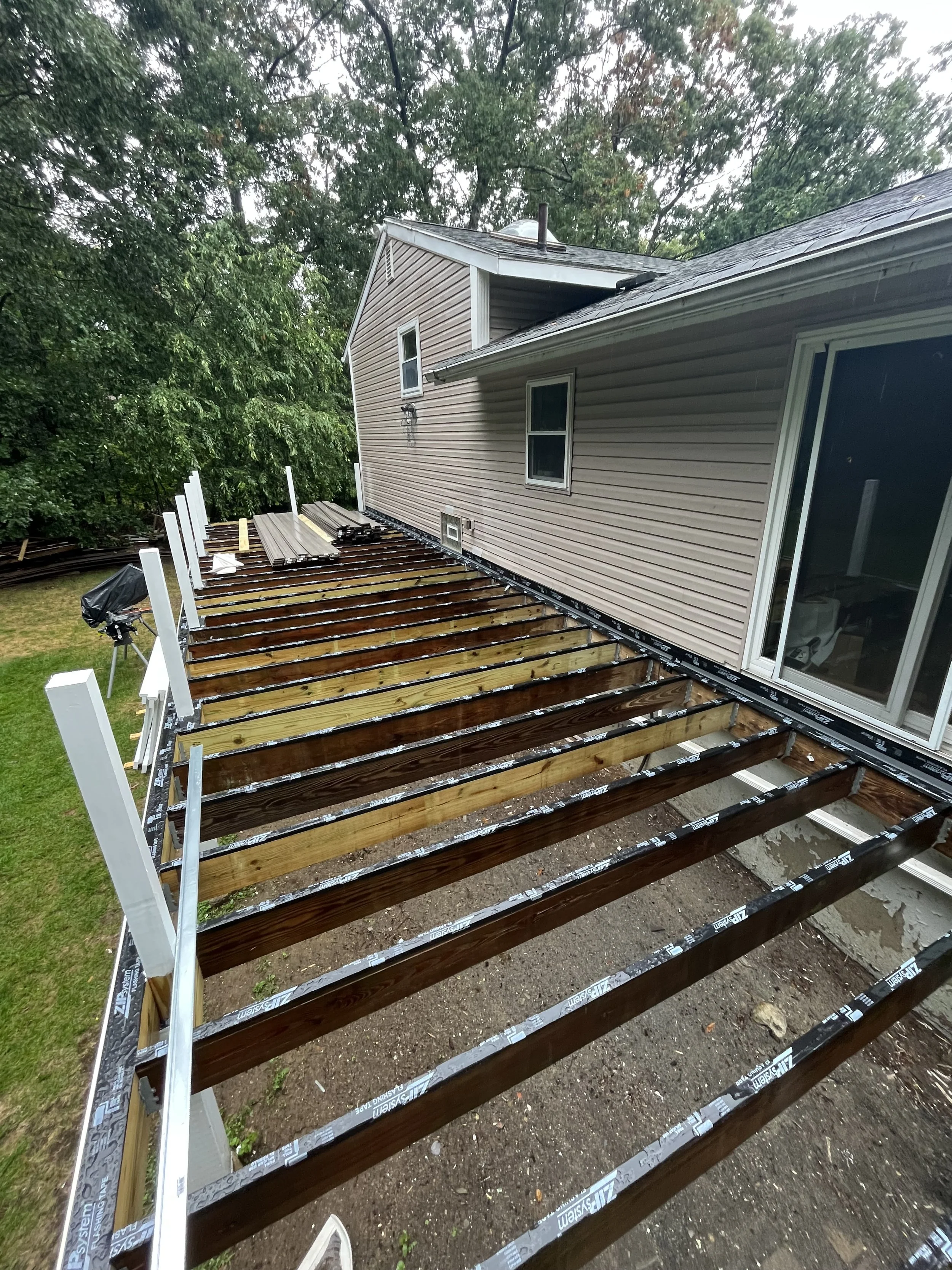A house with a deck under construction shows wooden beams and support posts installed on the frame. 