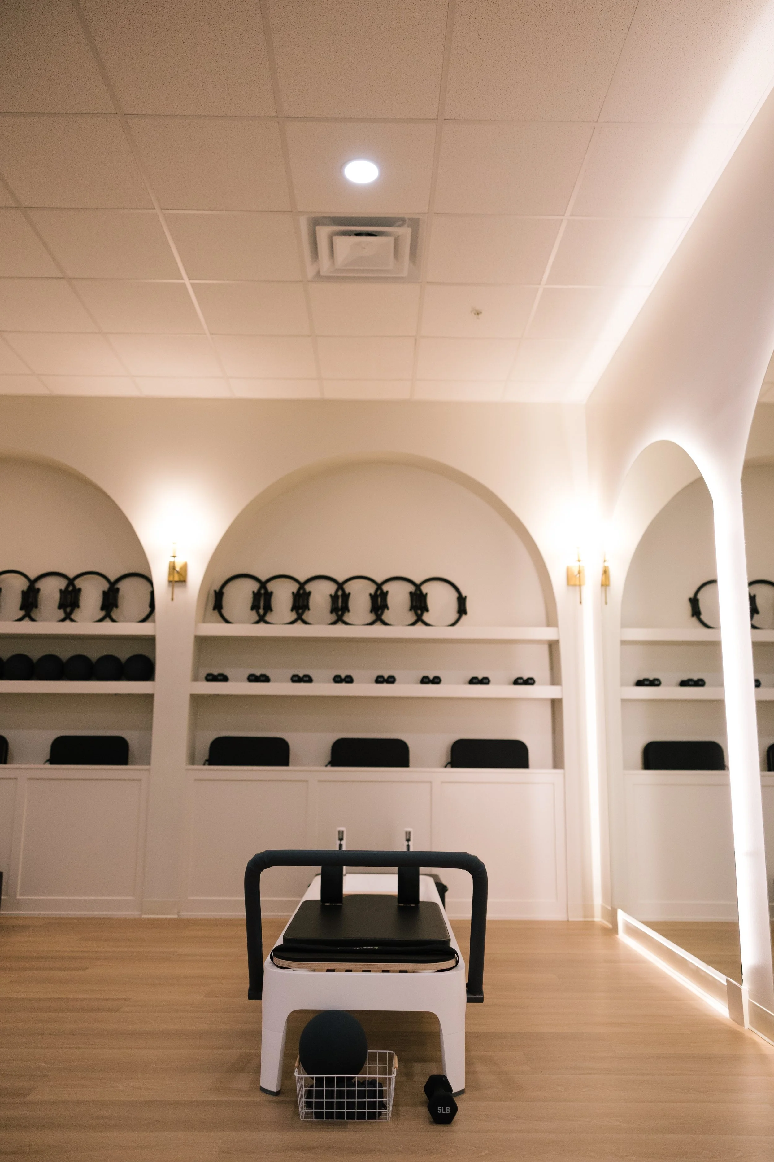 Fitness studio with black exercise equipment, white walls, and wooden floor, featuring a balance bench, a small basket with a black ball, and a 5 lb weight.