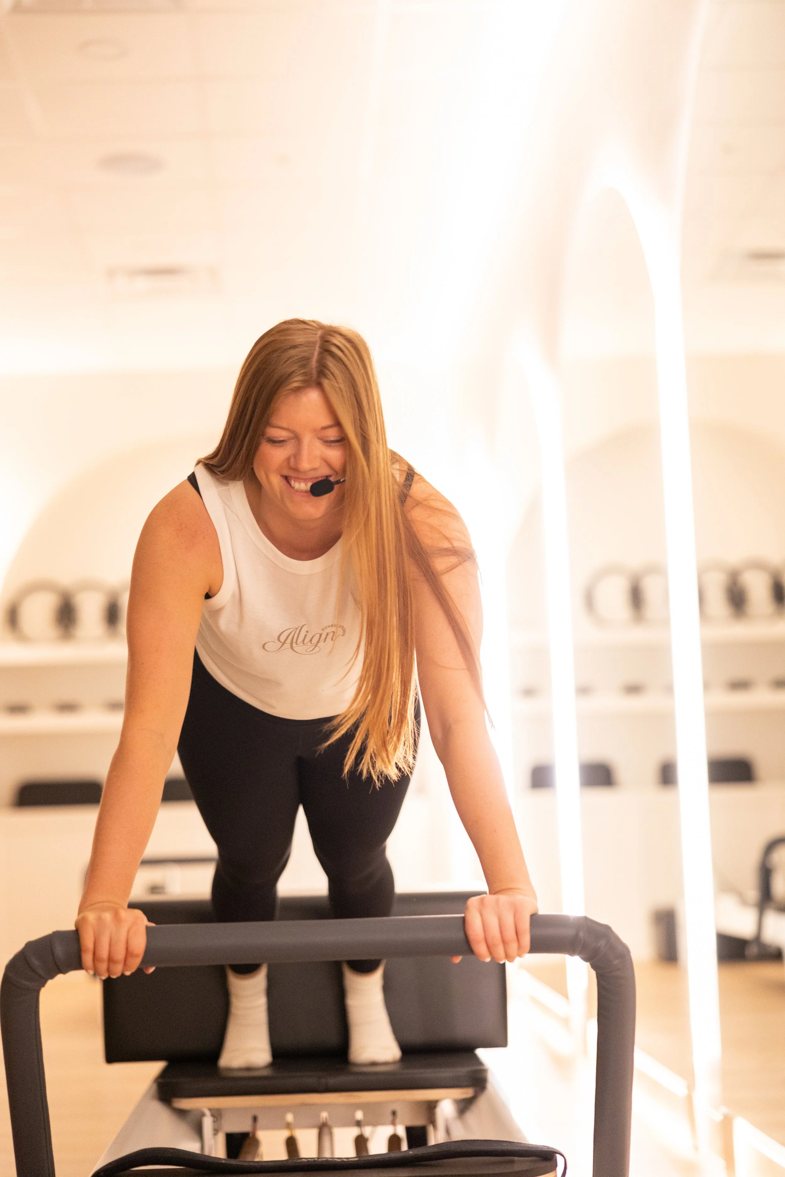 A woman with long red hair smiling and wearing a white tank top with 'Align' written on it, standing on a reformer in a fitness or Pilates studio, with a microphone headset.