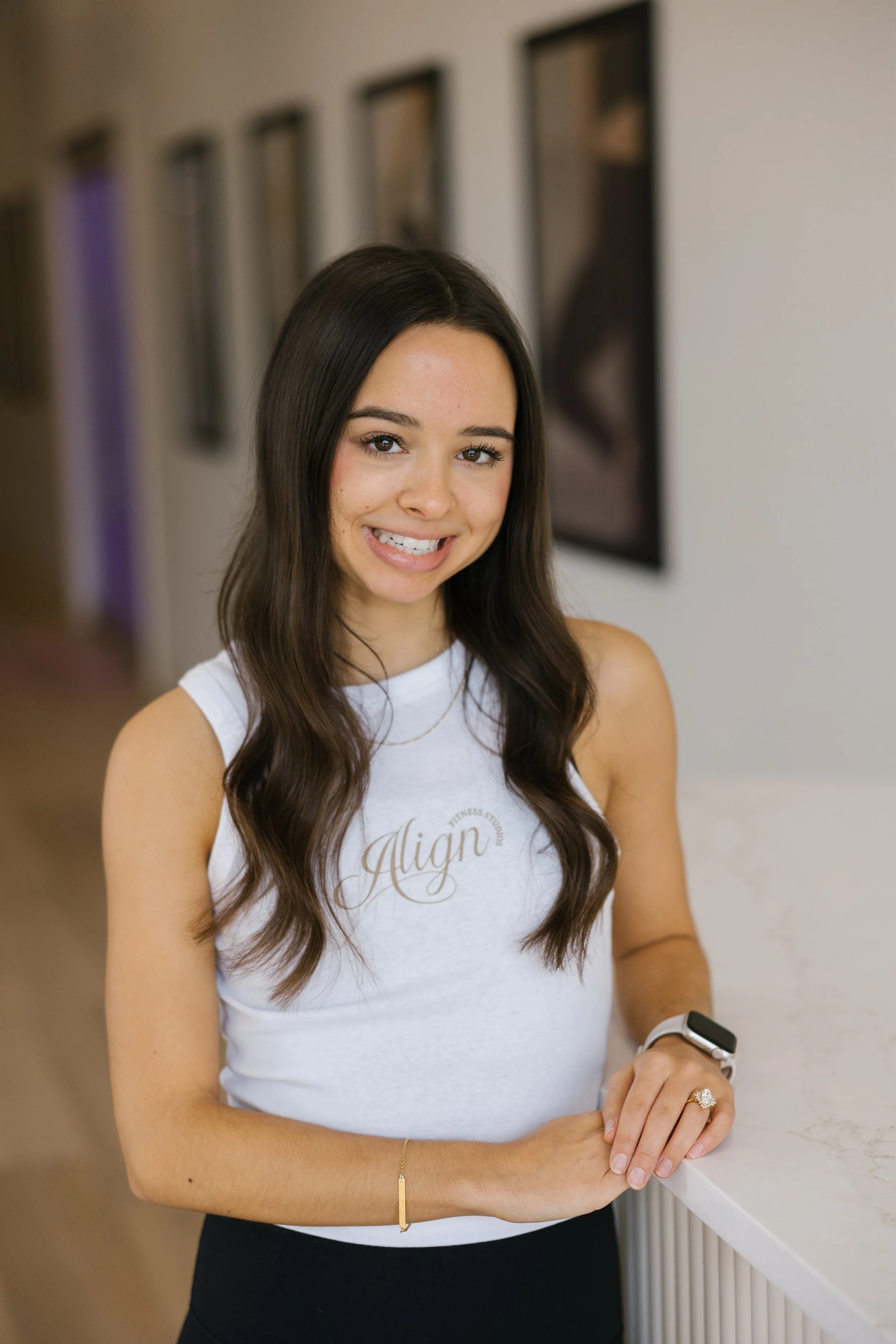 Young woman with long brown hair smiling, standing at a white marble counter in a modern interior, wearing a white tank top with 'Align' written on it, a smartwatch, and a ring.
