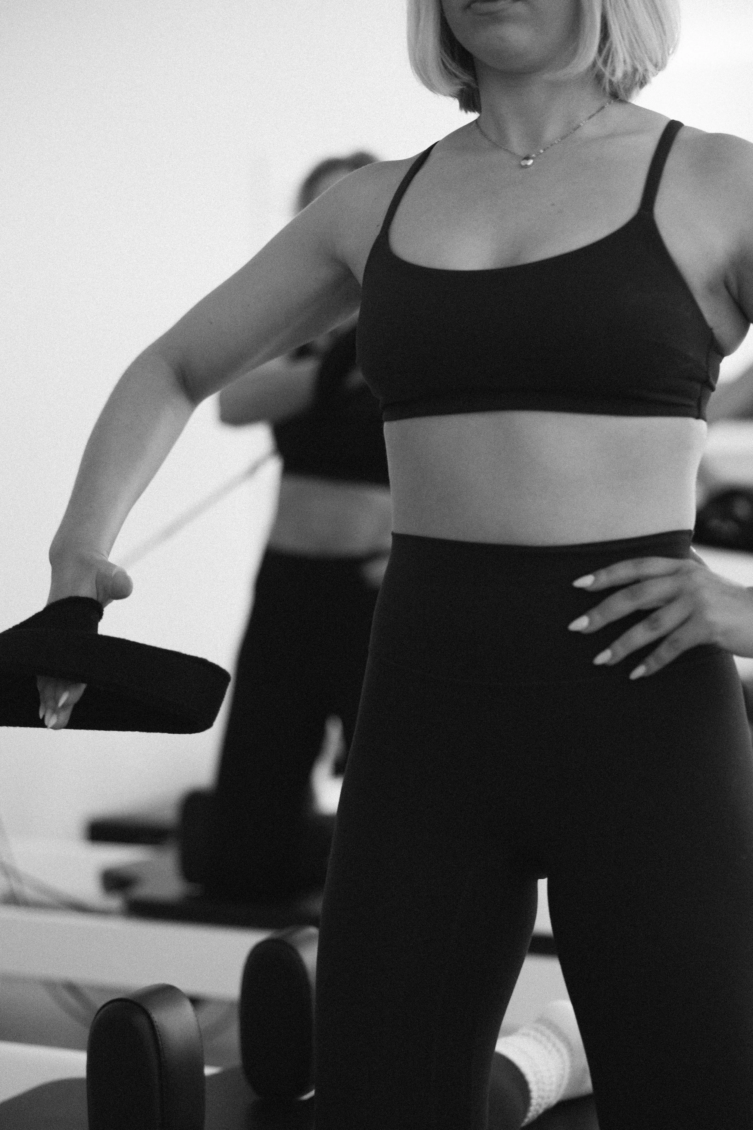 Woman holding weight in gym, wearing athletic clothing, with a blurred background of gym equipment and another person exercising.