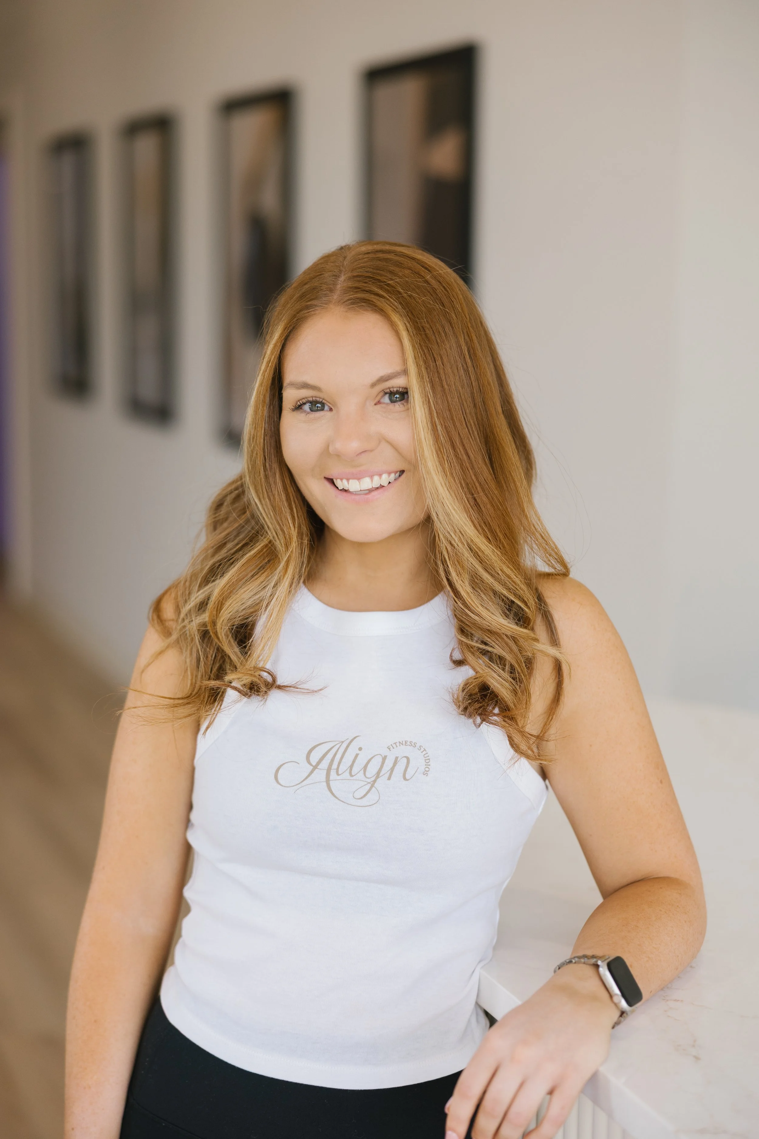 A young woman with long red hair, wearing a white sleeveless shirt with 'Align' written on it, smiling at the camera, standing in a well-lit room with framed pictures on the wall.