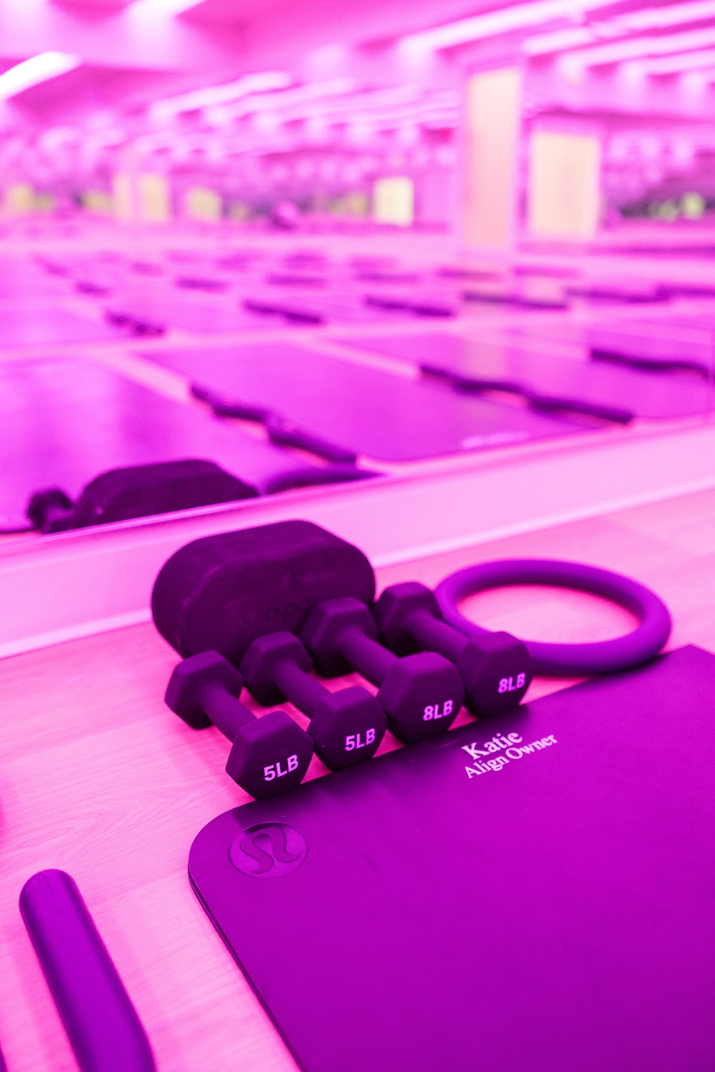 A close-up view of a workout area with a black mat labeled 'Katie Align Owner' and a set of small black dumbbells labeled 5LB, 8LB, on a wooden surface. Mirrors and purple lighting are visible in the background.