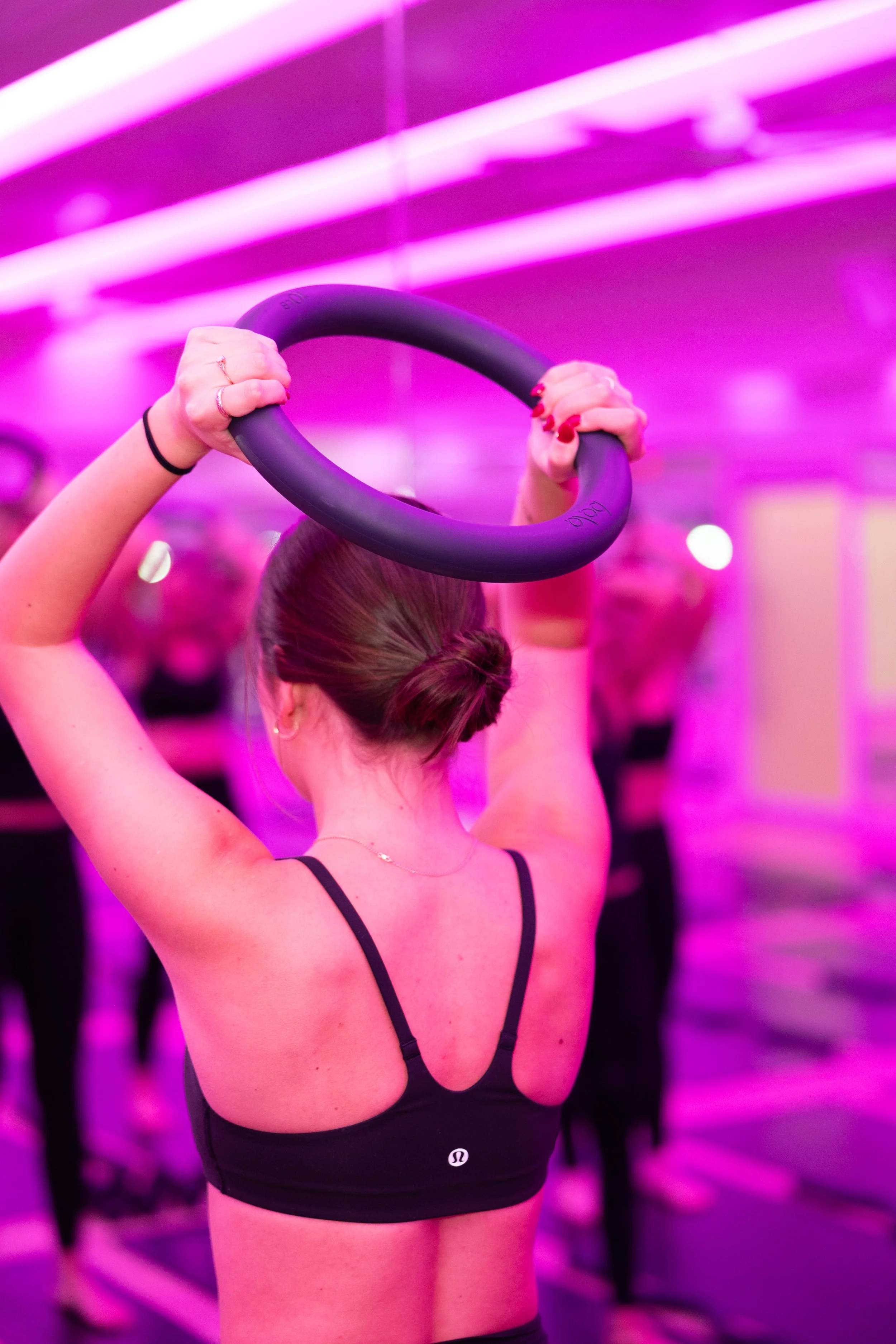 Back of woman holding a black fitness ring in a neon-lit fitness studio with purple and pink lighting.