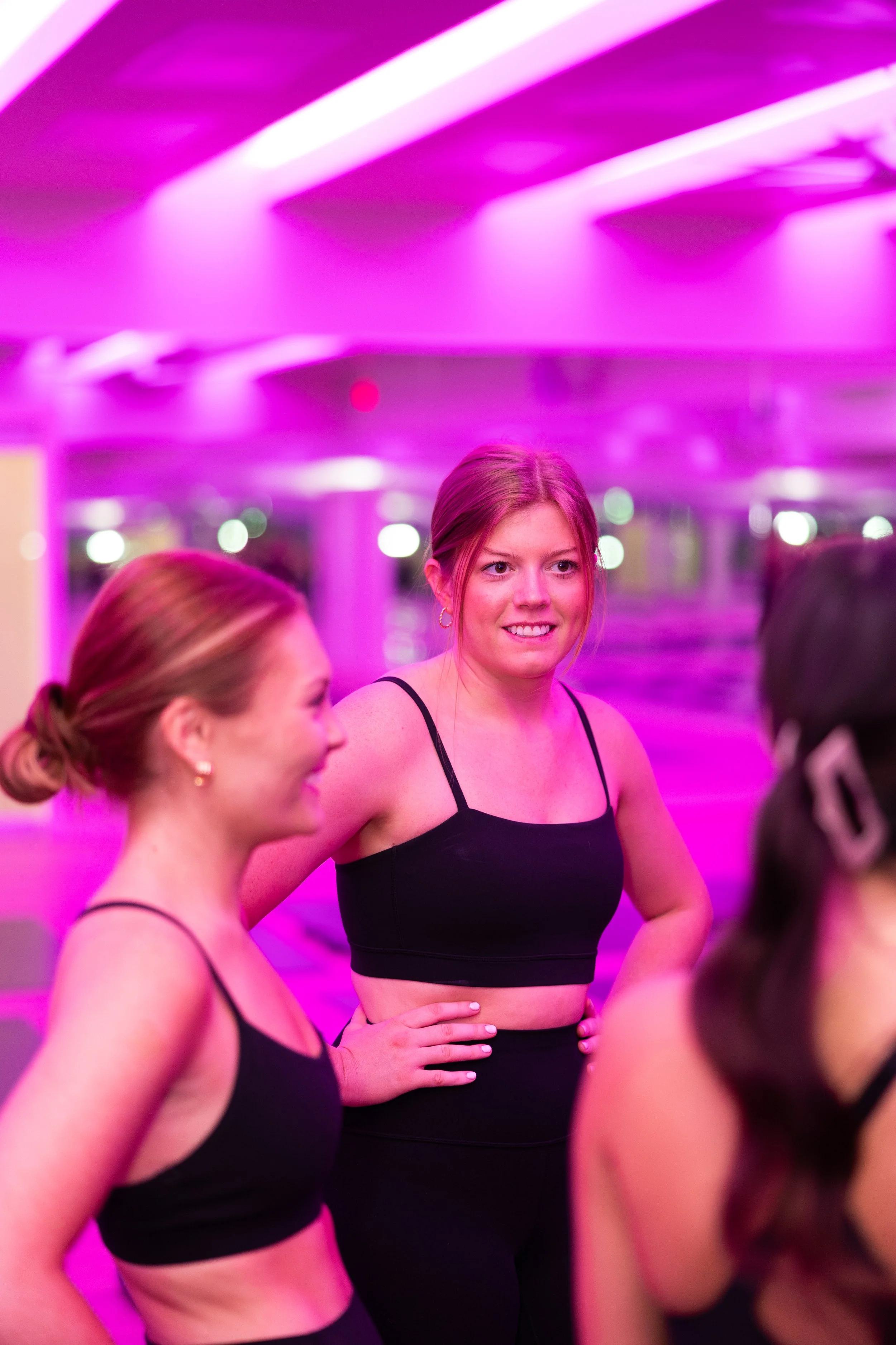 Three women in black activewear standing and talking in a room with purple lighting.