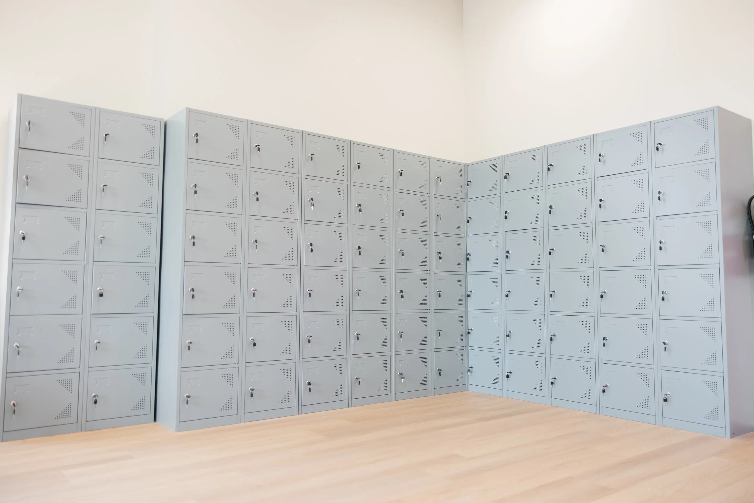 A room with many grey lockers with locks, arranged in a U-shape against white walls, with a light wood floor.