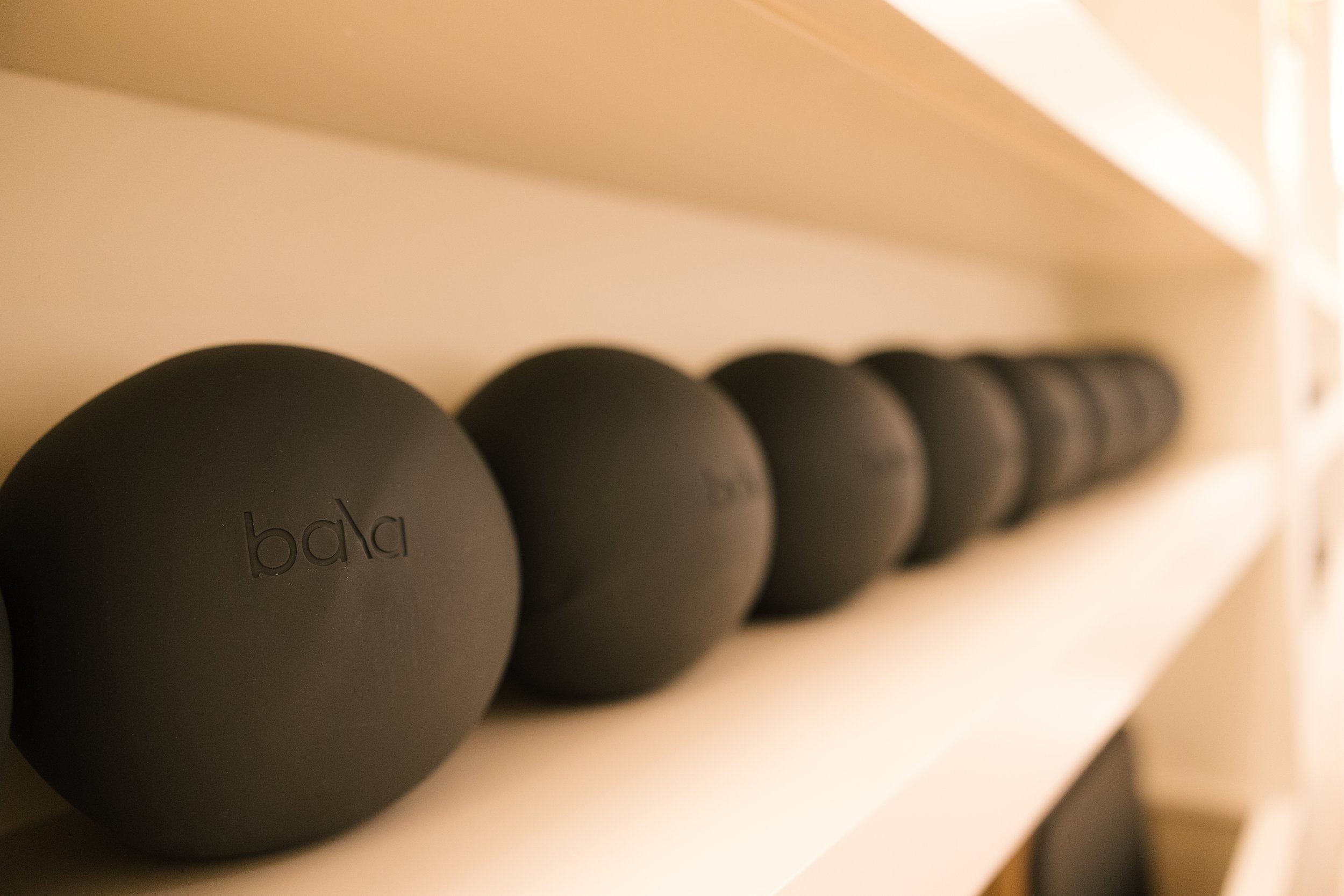 A row of black dumbbells on a white shelf, with the front dumbbell labeled "bala" and the others decreasing in size, against a beige background.