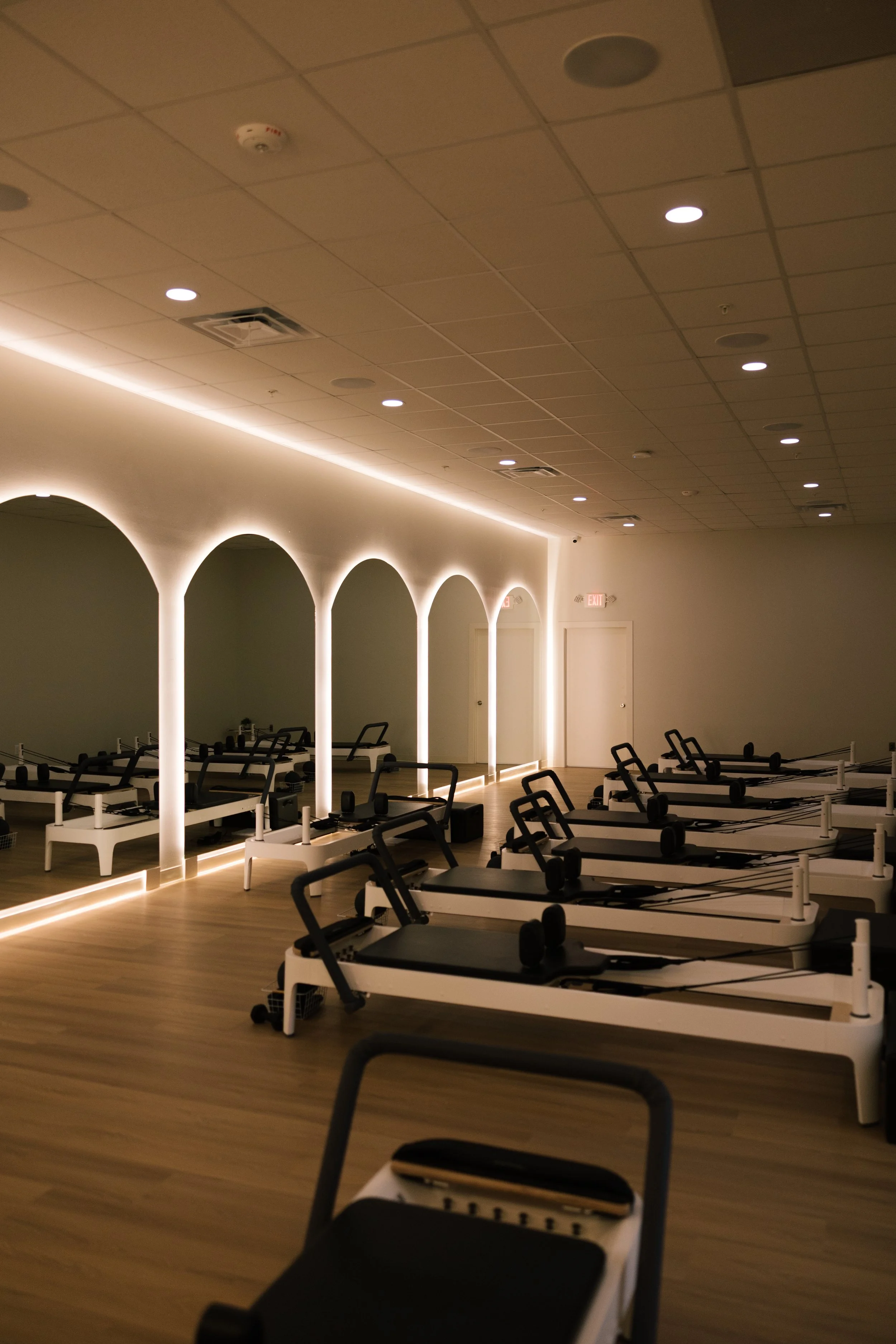 A Pilates or yoga studio with black reformer machines arranged in rows, illuminated by warm ambient lighting and a series of arch-shaped wall lights along one side of the room.