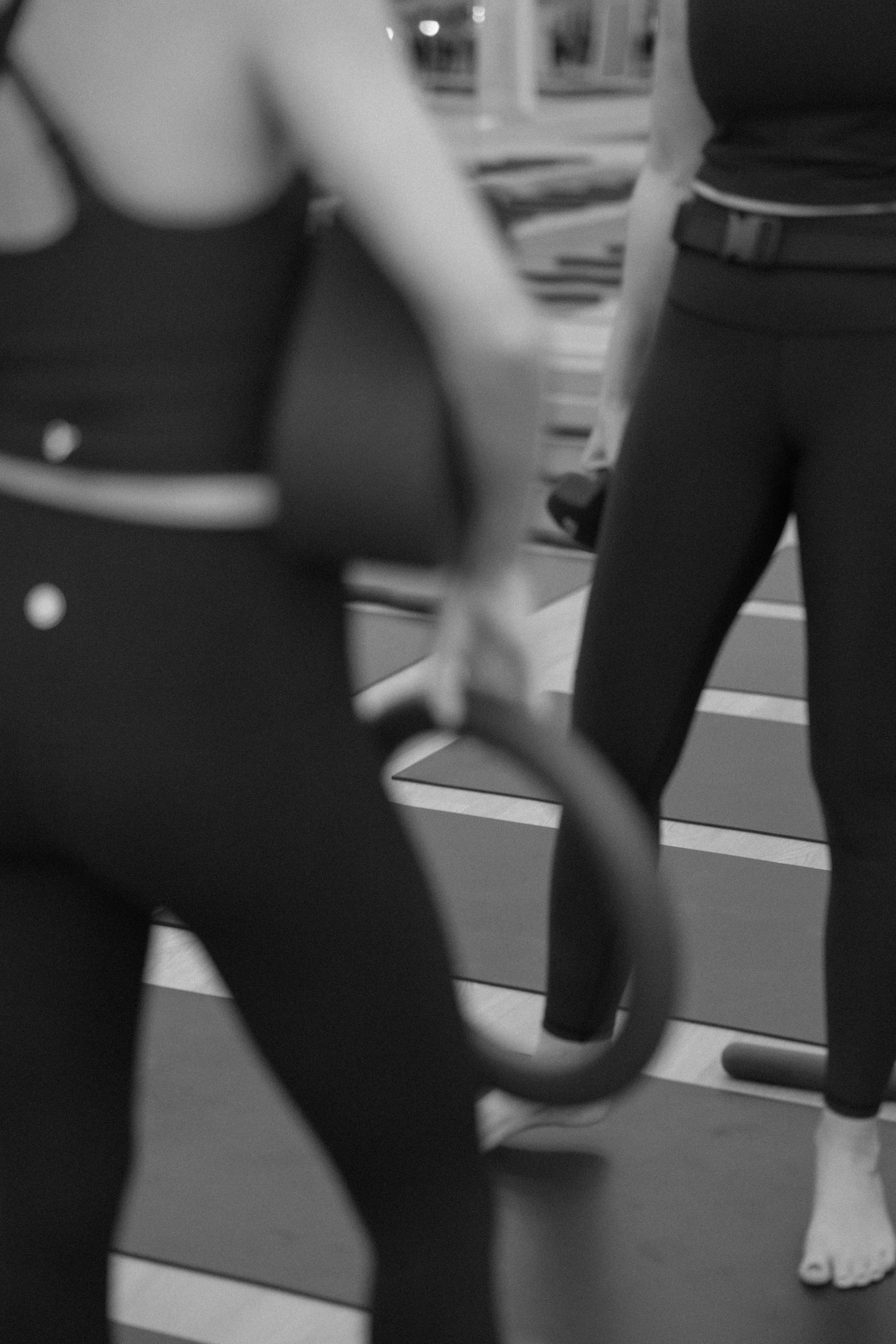 Two women practicing yoga or stretching, holding hands, in an indoor gym or studio with mats on the floor.