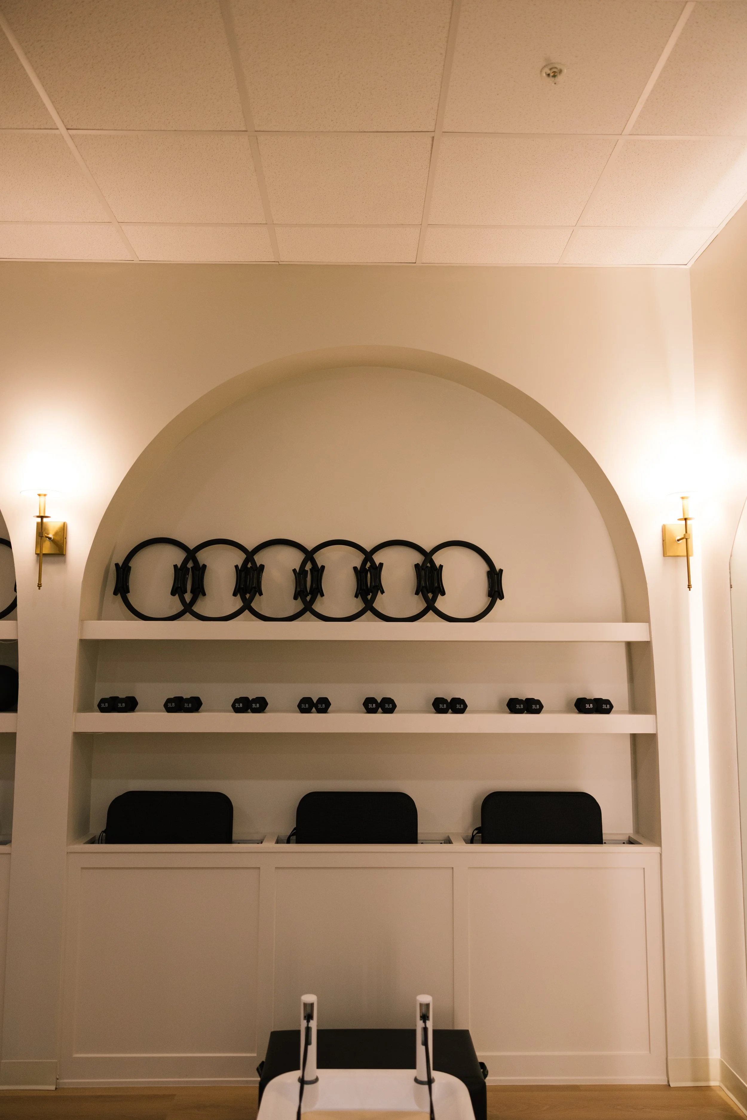 A white wall with an arched decorative feature, shelves holding exercise equipment, and wall sconces providing lighting.