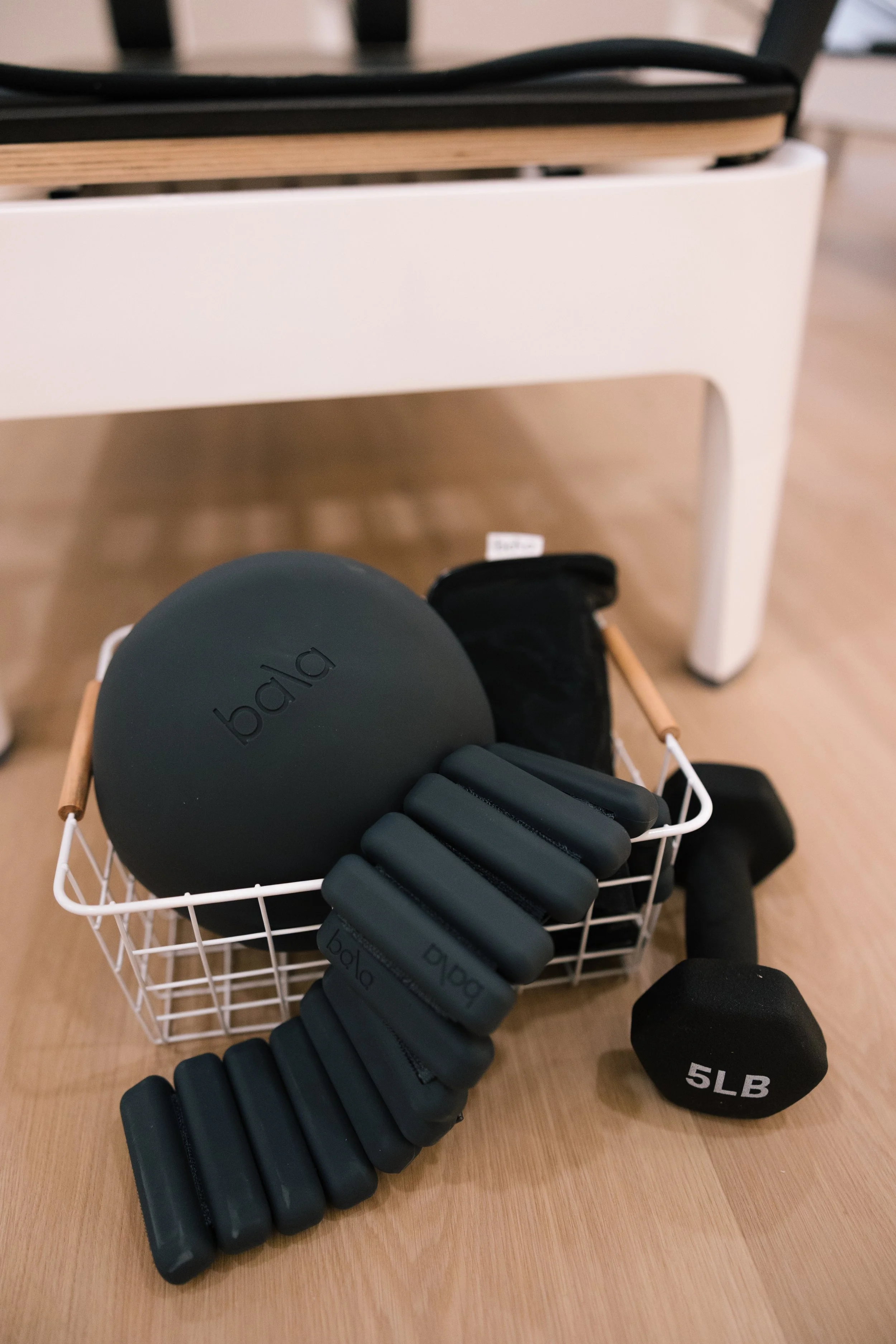 Home workout equipment including a black balance ball, black hand weights labeled 5LB, and a small black pouch, all placed in a white basket on a wooden floor.