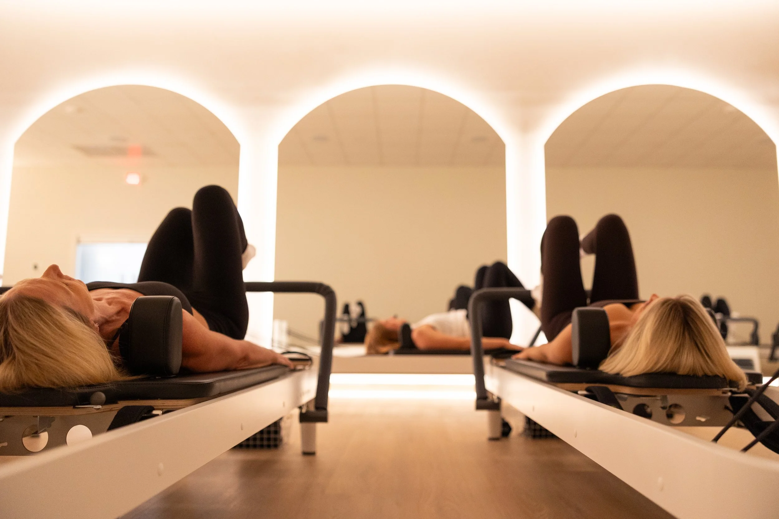 People lying on exercise mats during a fitness or stretch class in a dimly lit room with arched ceilings and soft backlighting.