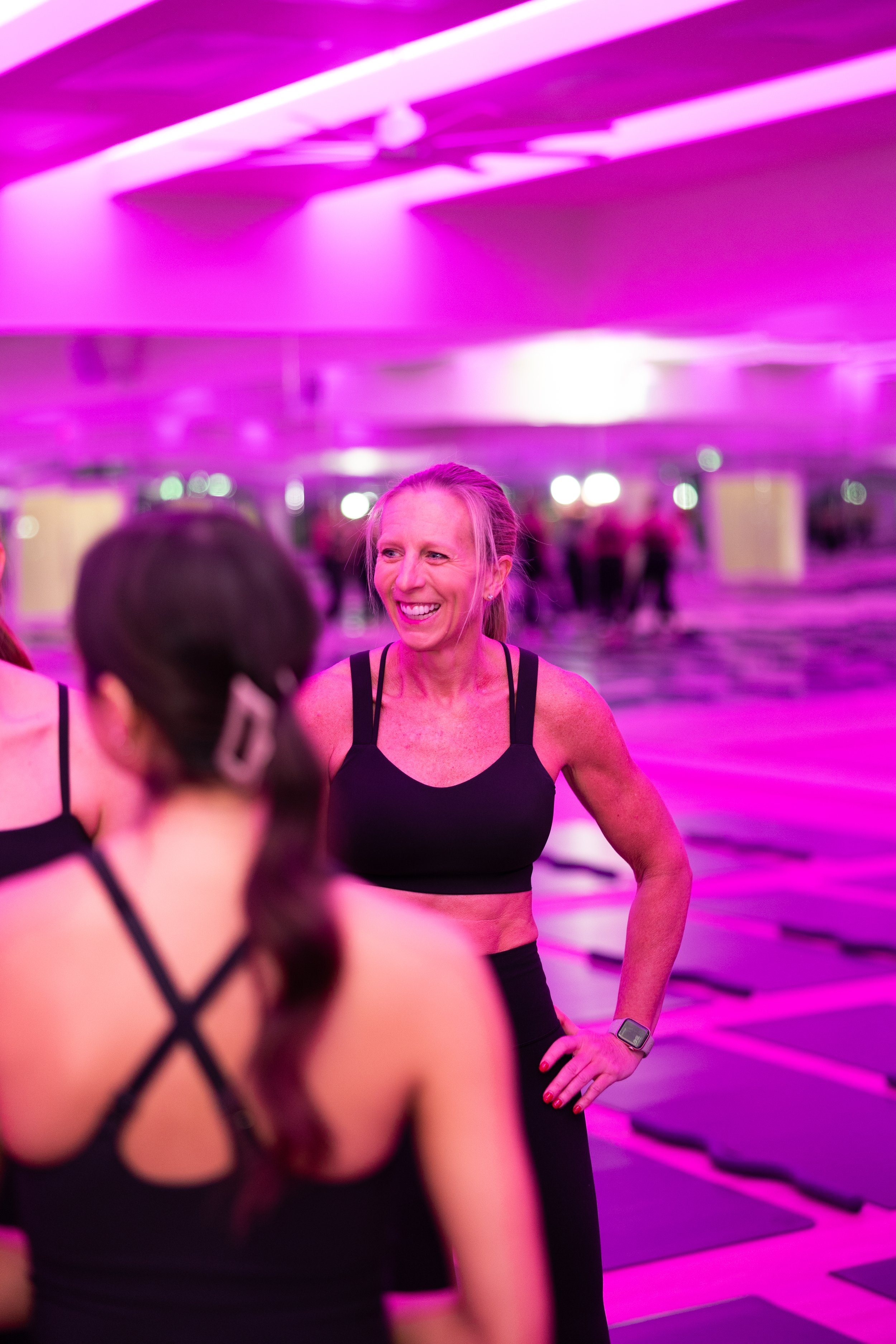Women in workout clothes talking in a fitness class with pink lighting and exercise mats on the floor.