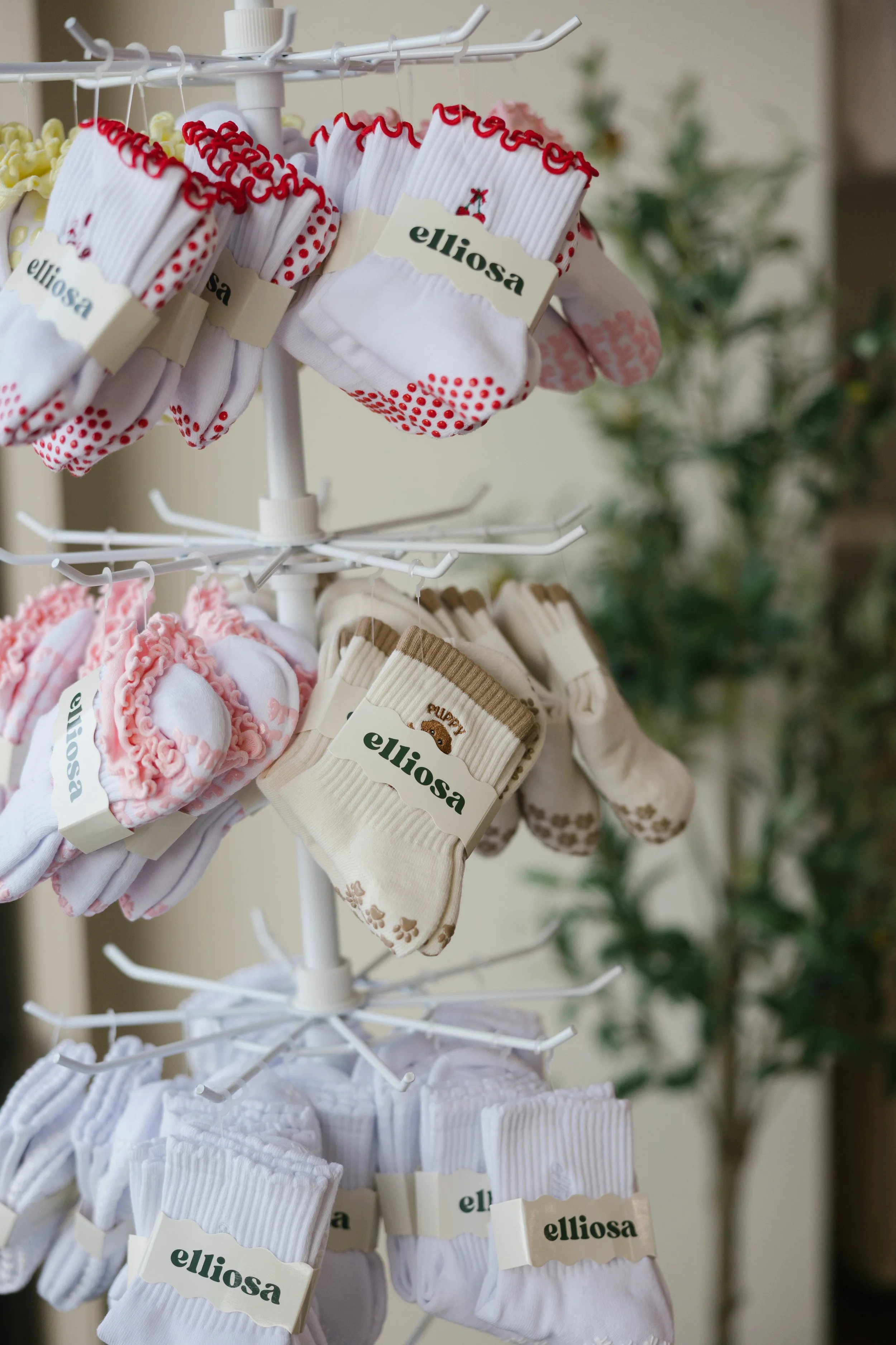 A display stand with multiple pairs of baby socks in various colors and patterns, branded "elliosa", arranged on a rotating rack.