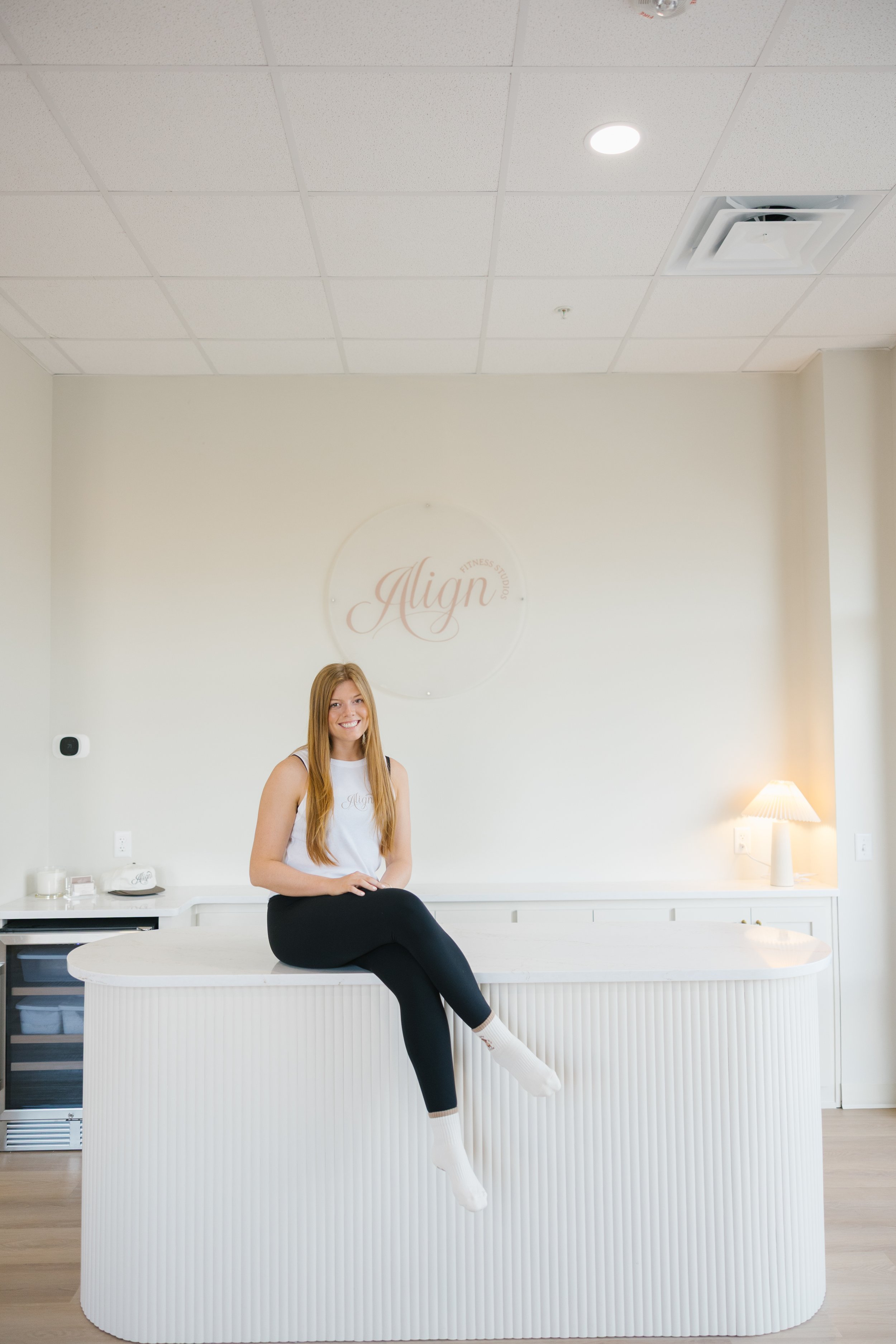 A woman with long red hair sitting on a white reception desk in a fitness studio lobby, smiling, with a sign that reads 'Align' on the wall behind her, and a lamp on the right side of the counter.
