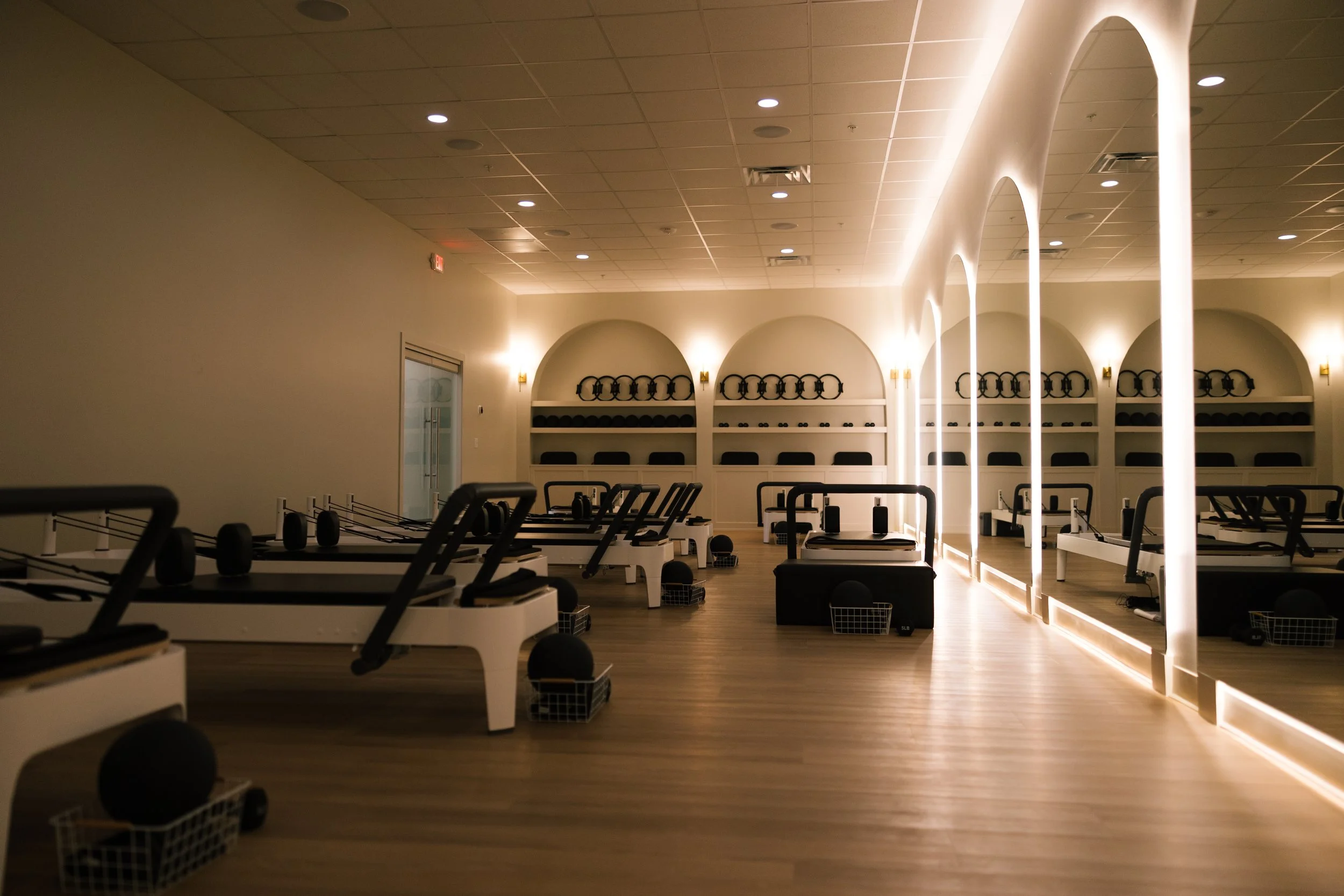 A Pilates or fitness studio with reformer machines arranged in rows, illuminated by soft ambient lighting, with shelves containing exercise equipment in arched alcoves at the back and a large mirror on the right wall.
