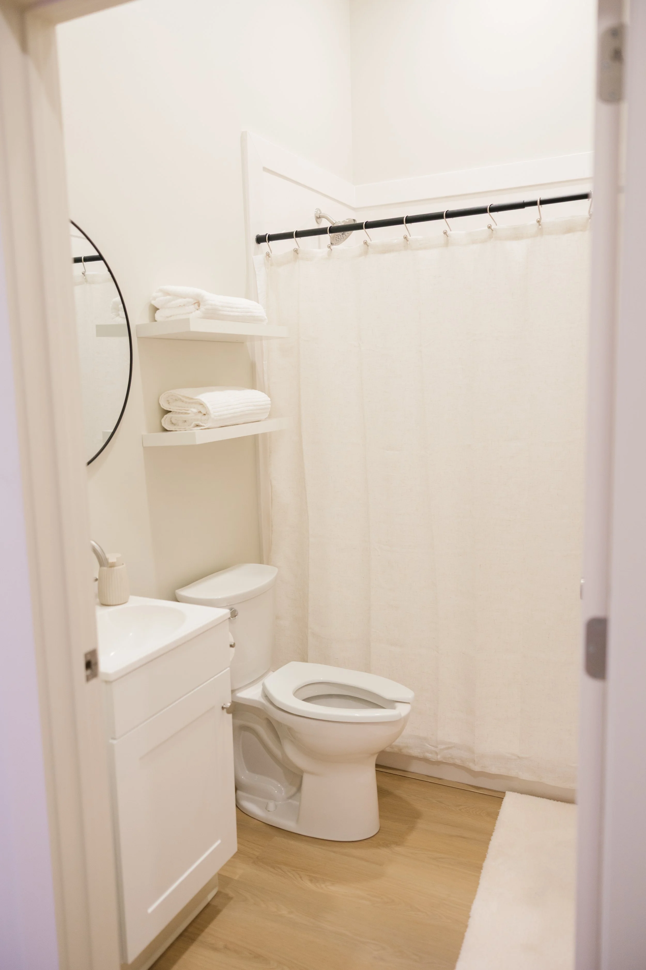 A small, minimalist bathroom with a white toilet, a white vanity with a sink, a small shelf with folded white towels, a round mirror, a shower with a curtain, and a light-colored wooden floor.