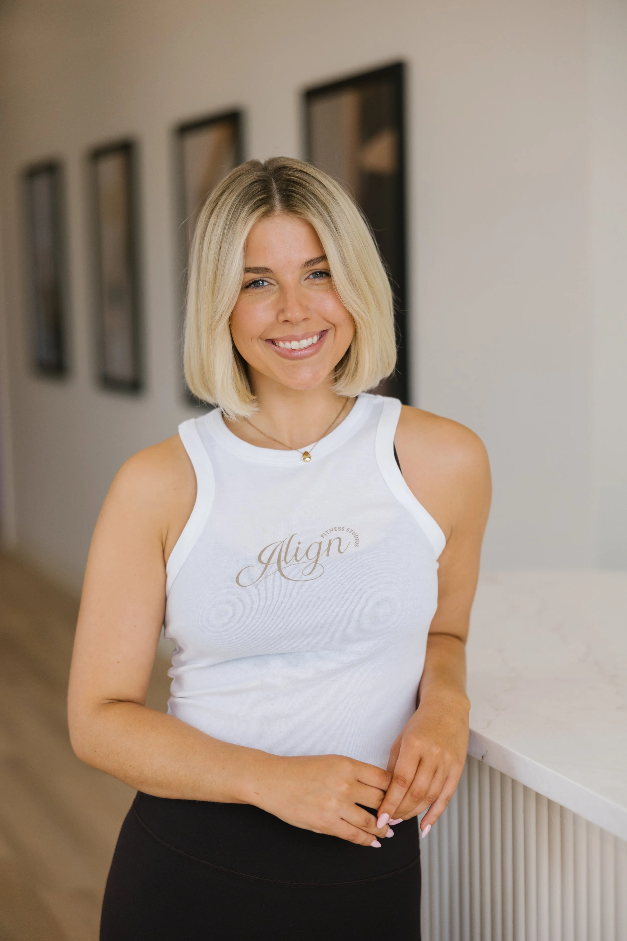 A smiling woman with blonde hair, wearing a white sleeveless top with 'Align' written on it, standing in a modern interior space.