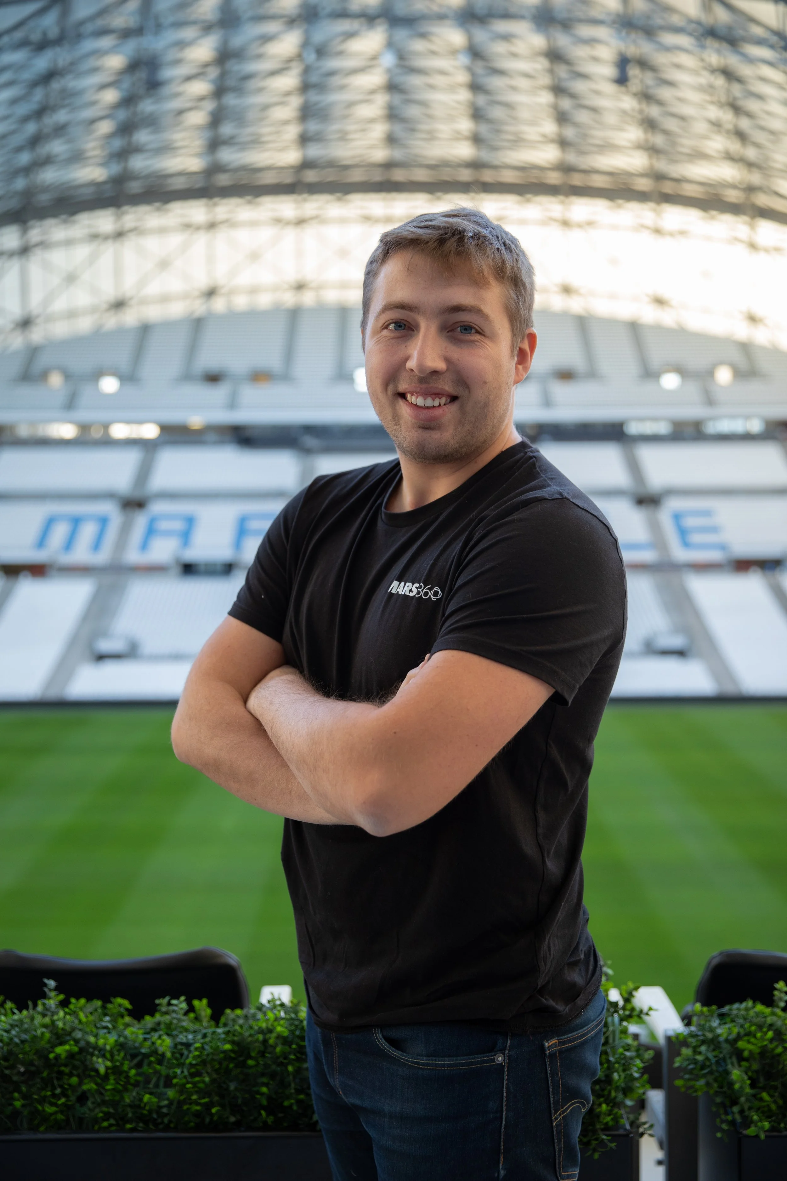 Jeune homme souriant avec un t-shirt noir, debout dans un stade vide, avec la pelouse verte en arrière-plan.
