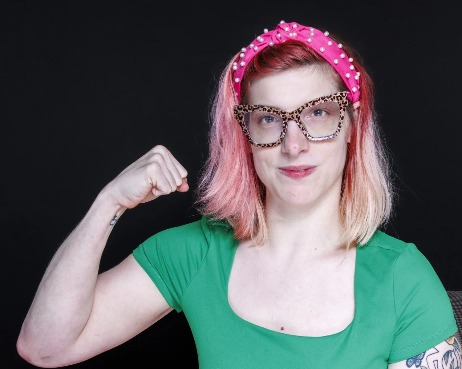 Myself flexing bicep, I am wearing a dark green short sleeve shirt. I have light pink, short hair, wearing a bright pink head band with white pearls and cheetah print glasses