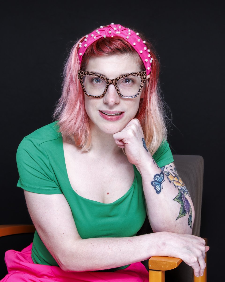 Kaylin, sitting in a chair my right arm is pointed toward my chin making a fist. I am wearing a dark green short-sleeved shirt. I have short light pink hair and wearing cheetah print glasses