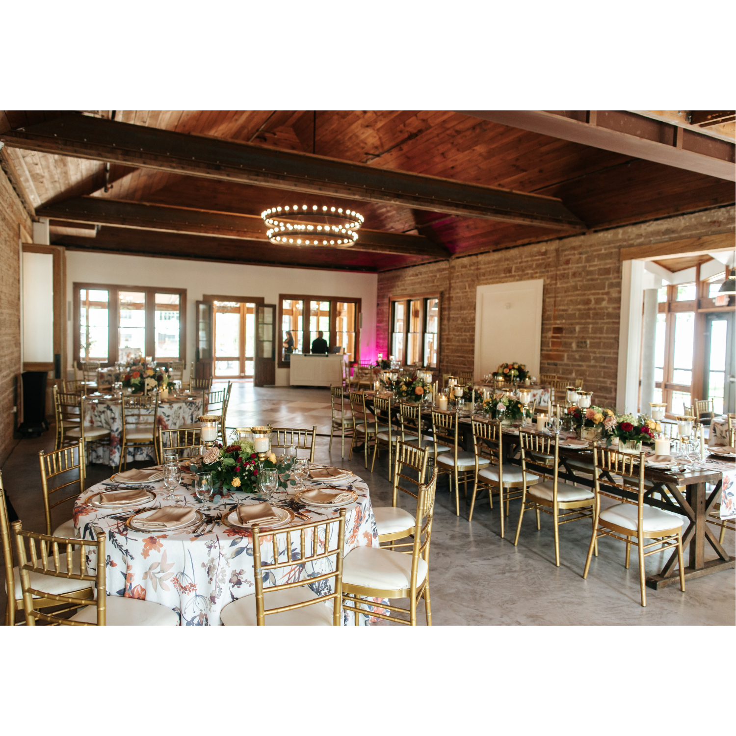Elegant event space decorated with floral centerpieces on tables adorned with candles, gold chairs, and floral tablecloth, with wooden walls and ceiling, illuminated by chandelier and natural light from windows.