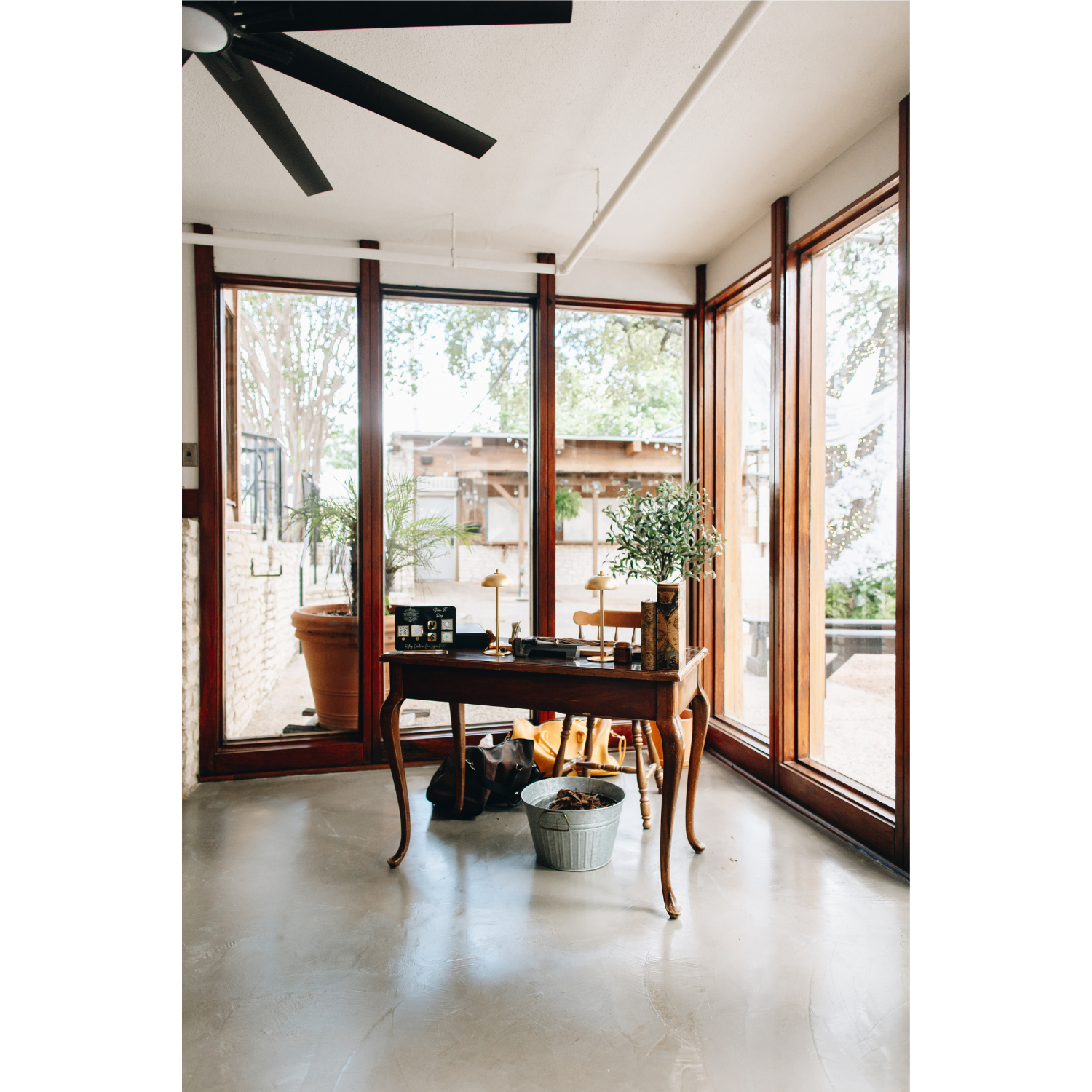 A cozy indoor space with large floor-to-ceiling windows showing an outdoor garden with trees. Inside, a small wooden table holds decorative items, a potted plant, and two small lamps. The floor is polished concrete, and a ceiling fan is partially vis