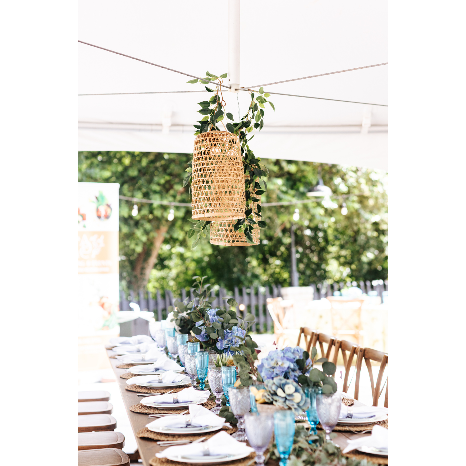 Long outdoor dining table decorated with floral arrangements, place settings, and colorful glassware, with hanging woven lanterns and string lights overhead.