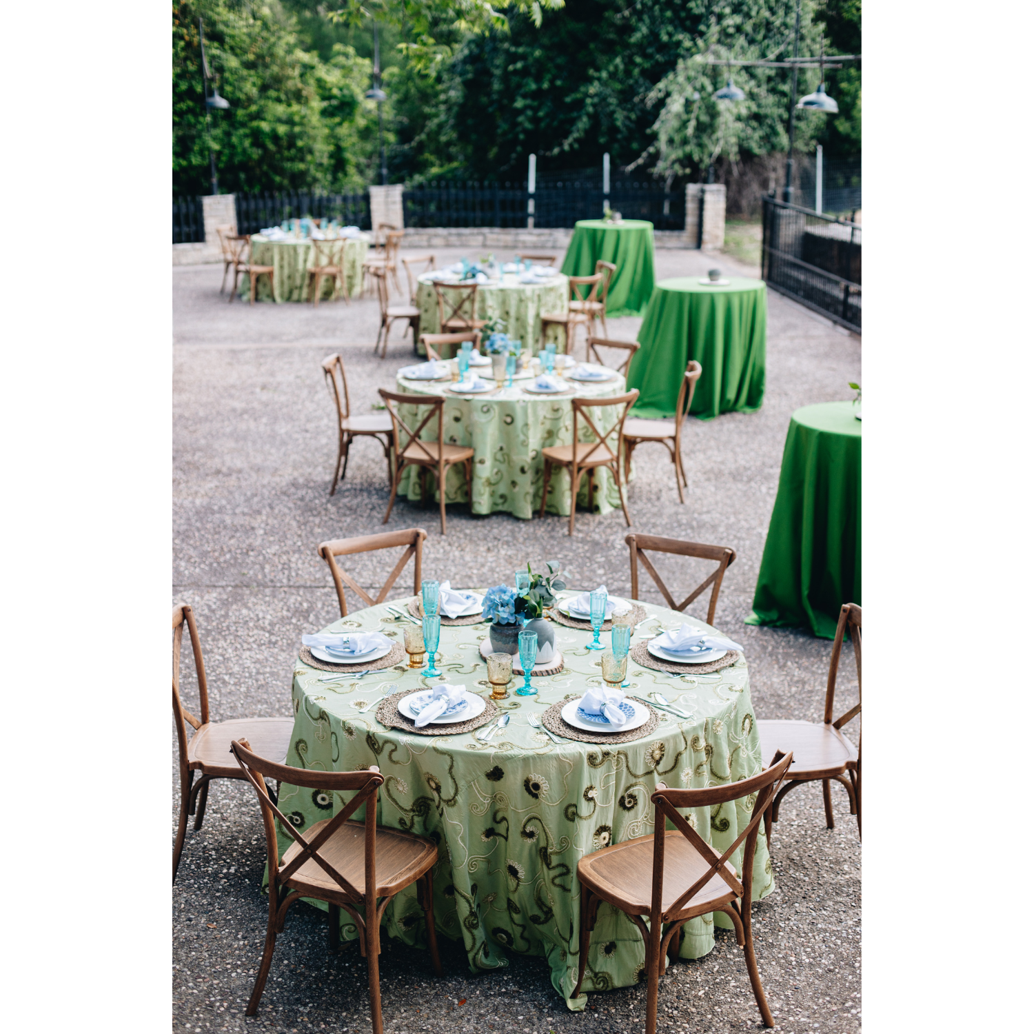 Outdoor banquet setup with round tables covered in floral green tablecloths, set with plates, napkins, silverware, and colorful glassware, surrounded by wooden chairs, on a gravel surface with trees and black fencing in the background.