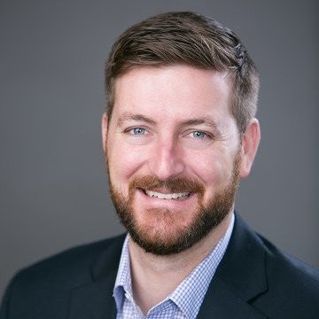 A professional headshot of a man with short brown hair, a beard, wearing a dark blazer and a light blue shirt, smiling against a gray background.