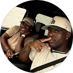Two young men sitting in a car, smiling, with one wearing a white cap and the other in a beige shirt.