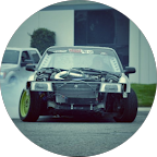 A race car on a track, partially disassembled with its hood open and front bumper missing, in a racing environment with other cars and trees in the background.