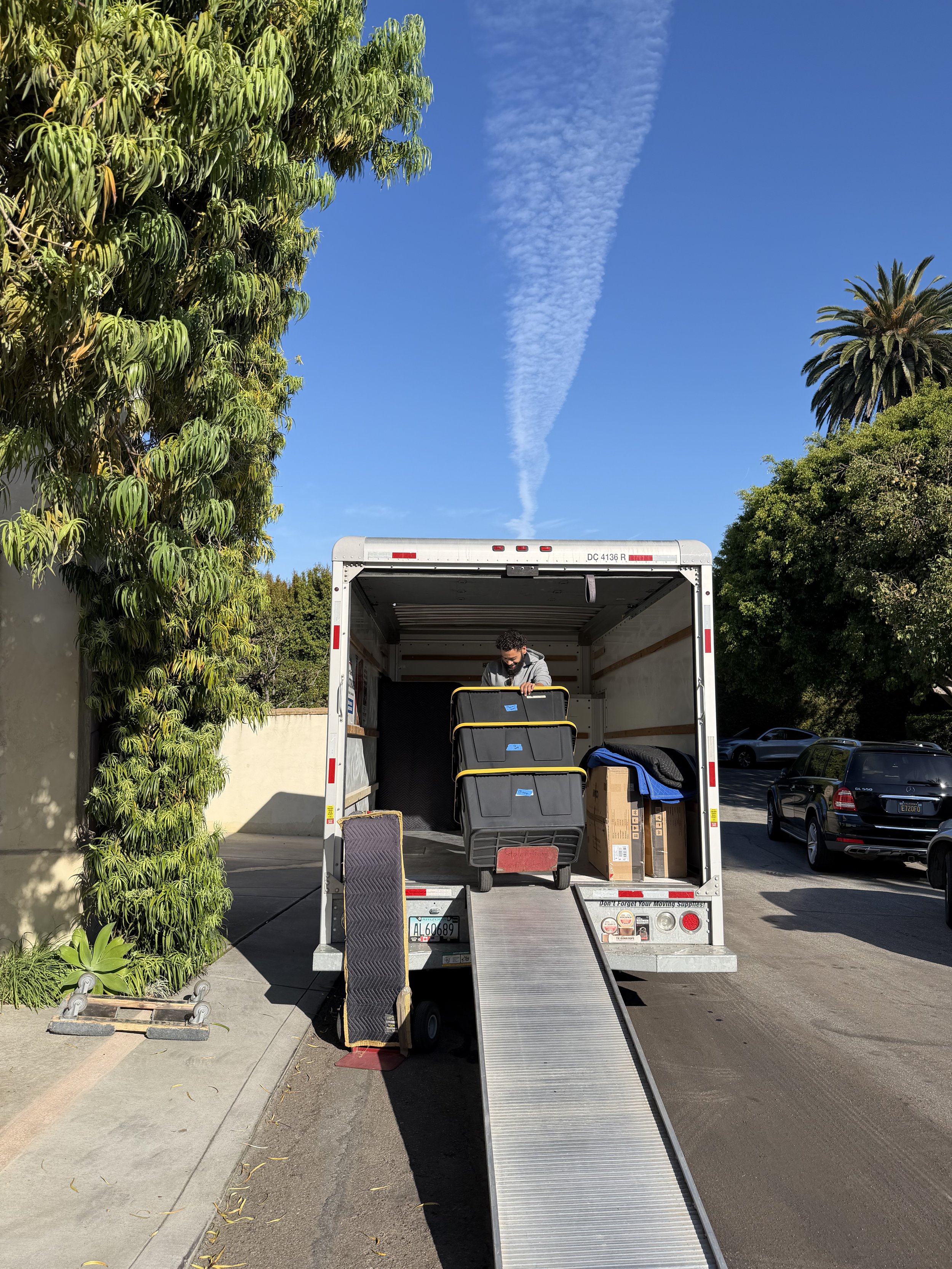 A man unloading black plastic storage containers from the back of a moving truck with a ramp, parked on a street next to green trees and several parked cars, under a blue sky with a contrail.