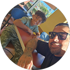 A man with a tattooed arm and sunglasses taking a selfie with a young boy at an amusement park or carnival, with a blue ship ride in the background.