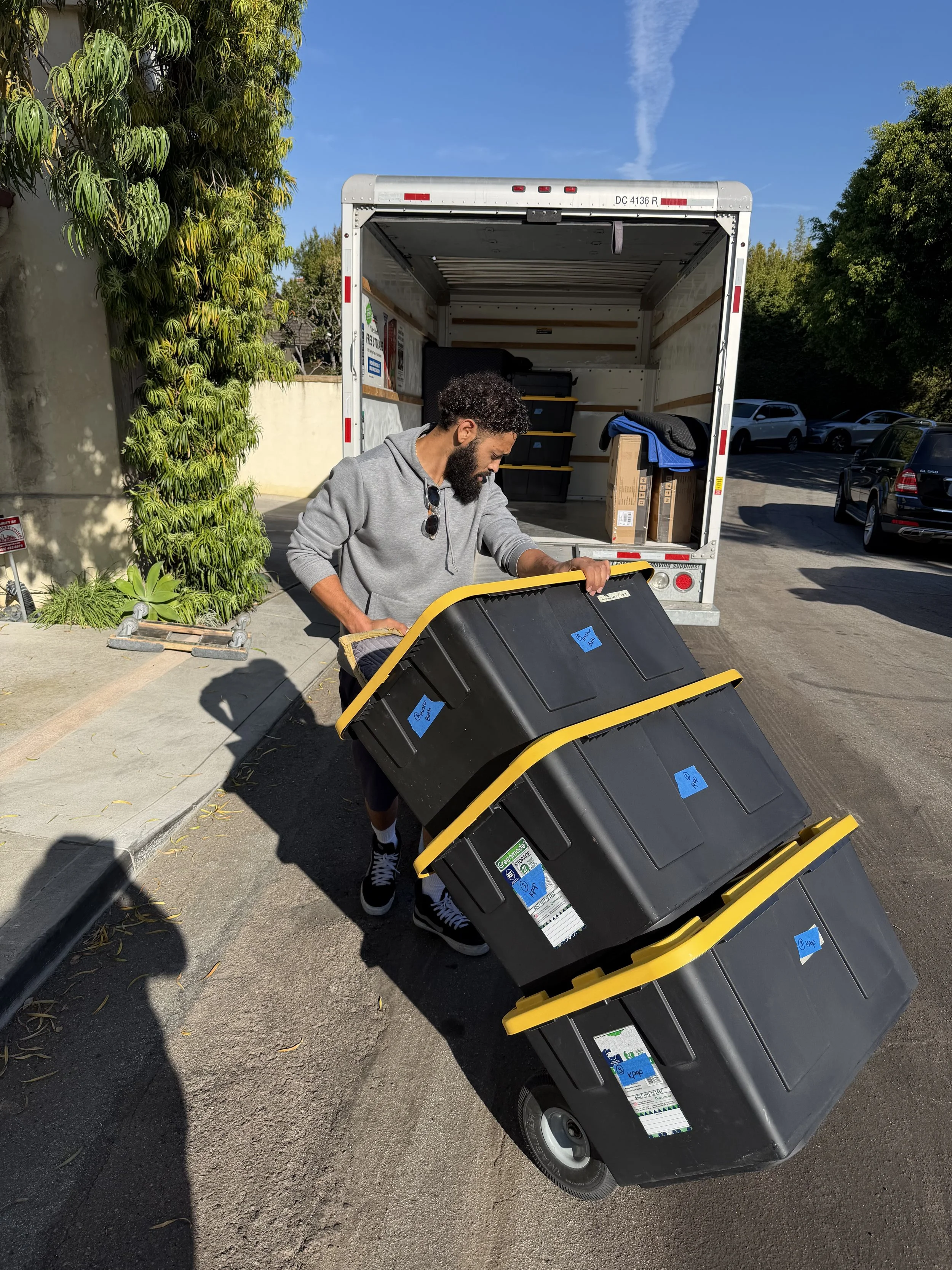 Man unloading black trash bins with yellow lids from a moving truck on a sunny day.