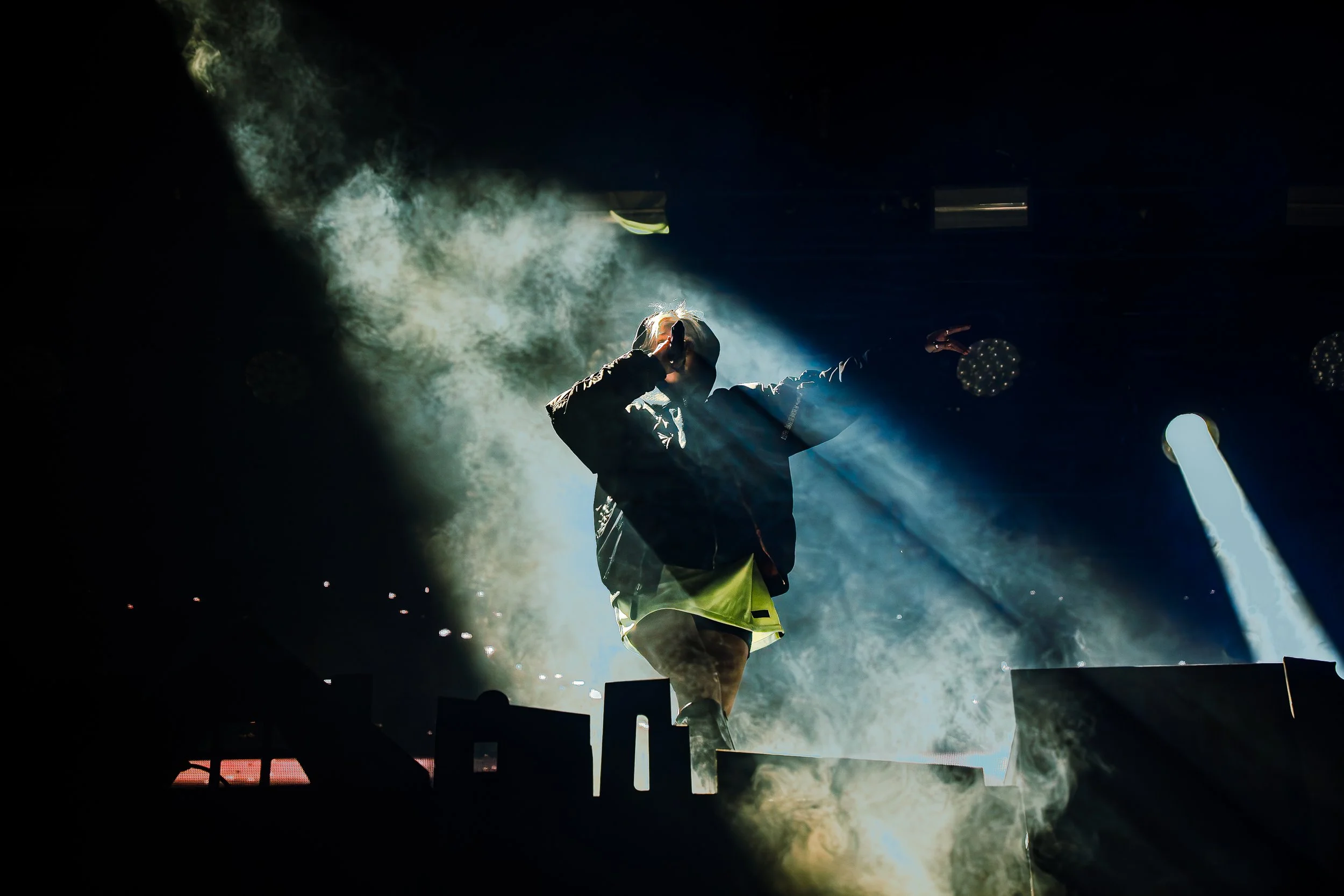 Ein Musiker auf einer dunklen Bühne singt in ein Mikrofon, beleuchtet von einem hellen Lichtstrahl, umgeben von Rauch oder Nebel.