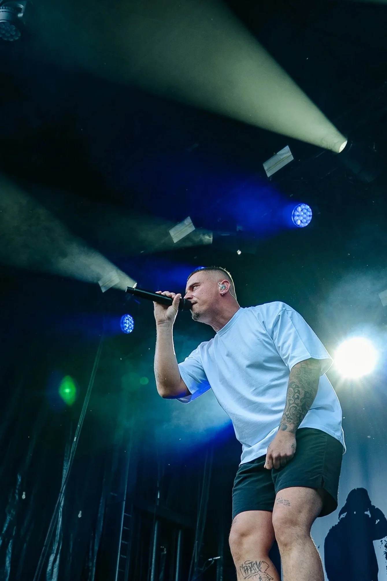 Ein Mann singt auf einer Bühne und hält ein Mikrofon in der Hand, er trägt ein weißes T-Shirt und schwarze Shorts, mit Tattoos an Armen und Beinen. Im Hintergrund sind Bühnenlichter und eine Person am DJ-Setup zu sehen.