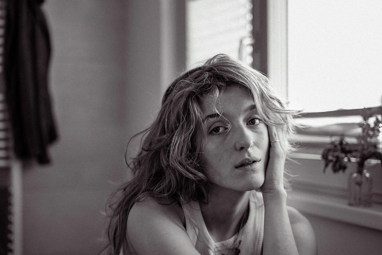Schwarz-weiß Fotografie einer jungen Frau mit lockigem Haar, die an einem Fenster sitzt.