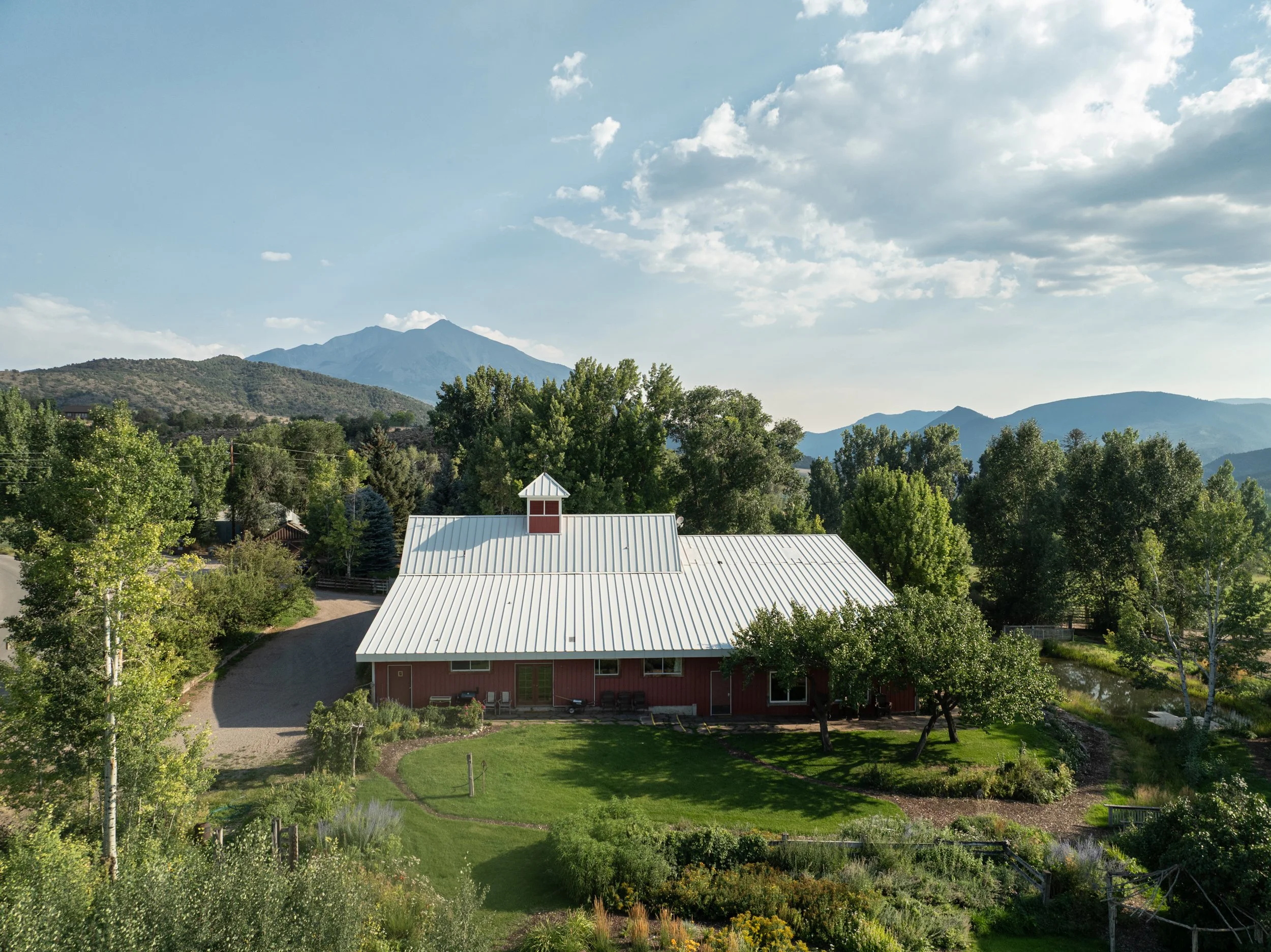 A large red barn with a metal roof and a small cupola, surrounded by lush green trees and a well-kept lawn, with mountains in the distance under a partly cloudy sky.